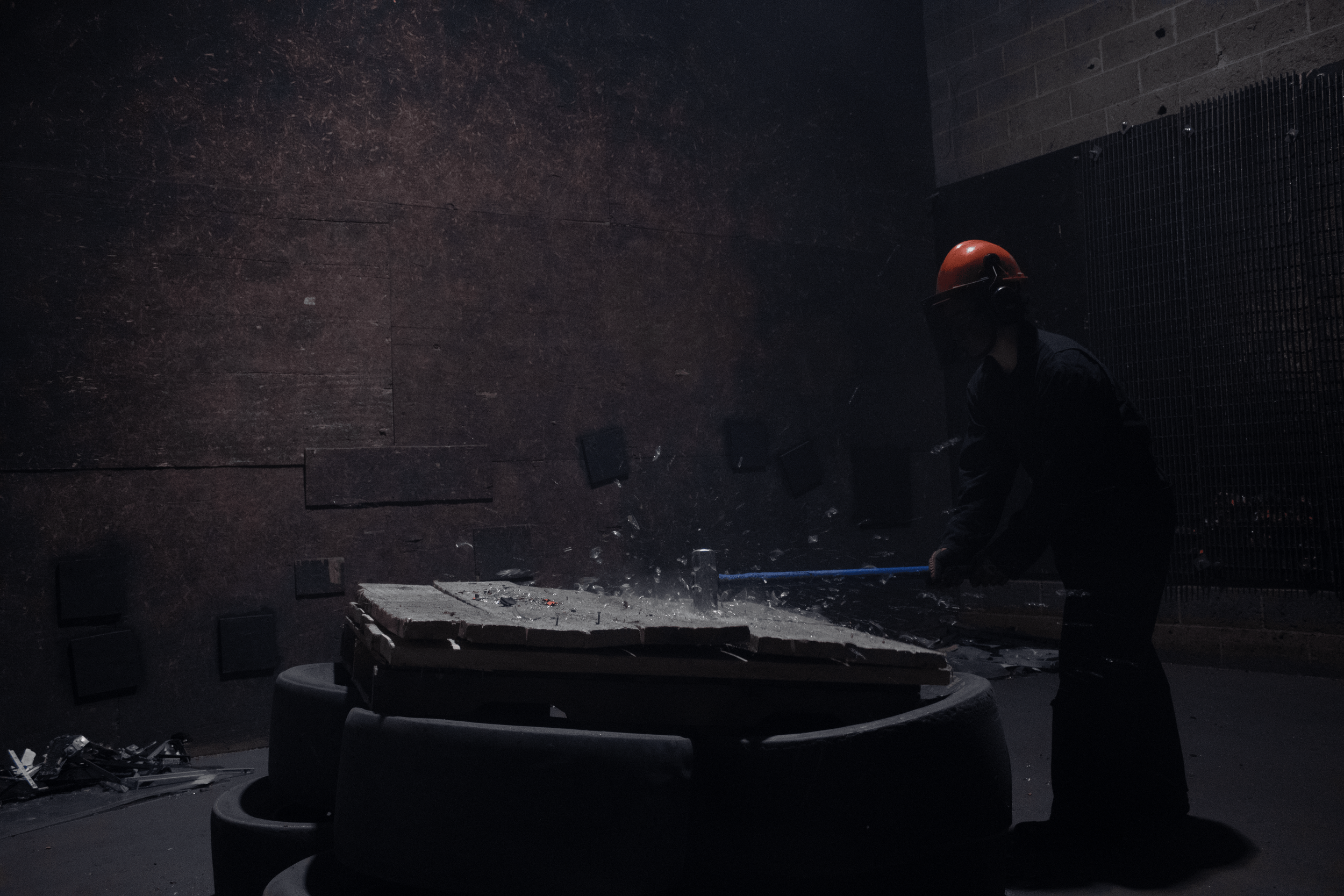 A person wearing a red safety helmet and face shield is smashing a piece of material with a crowbar, causing debris to fall onto the surface below in a dark industrial setting.