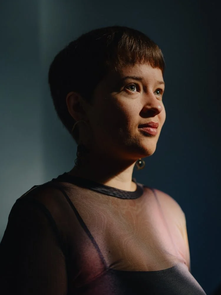 Portræt af en person med kort mørkt hår, iført gennemsigtigt sort top, stående i side, med blødt lys der fremhæver ansigtet i en mørk baggrund.