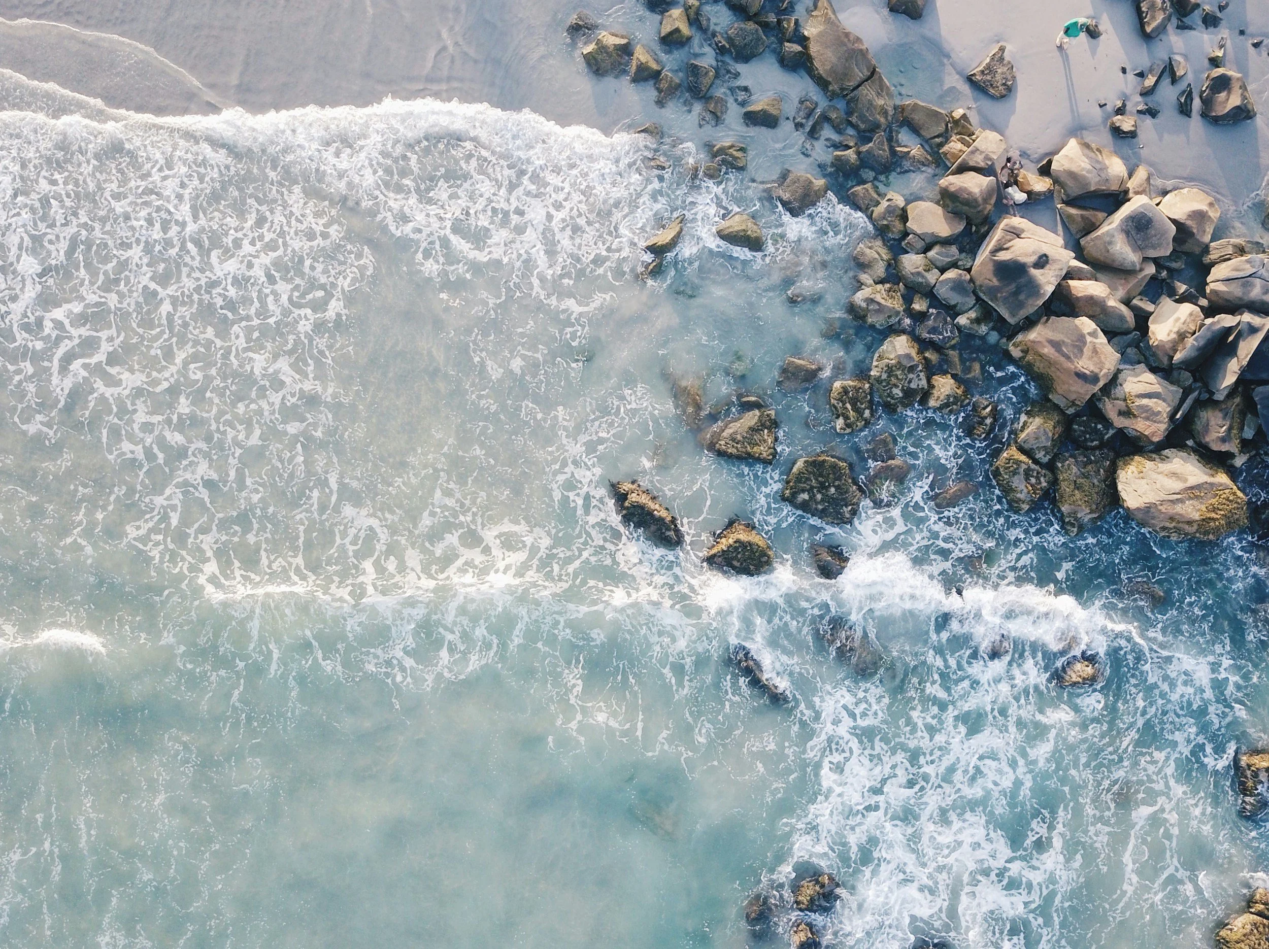 An aerial view of the shoreline showing ocean waves crashing onto a sandy beach with a large cluster of rocks and two people standing on the rocks.