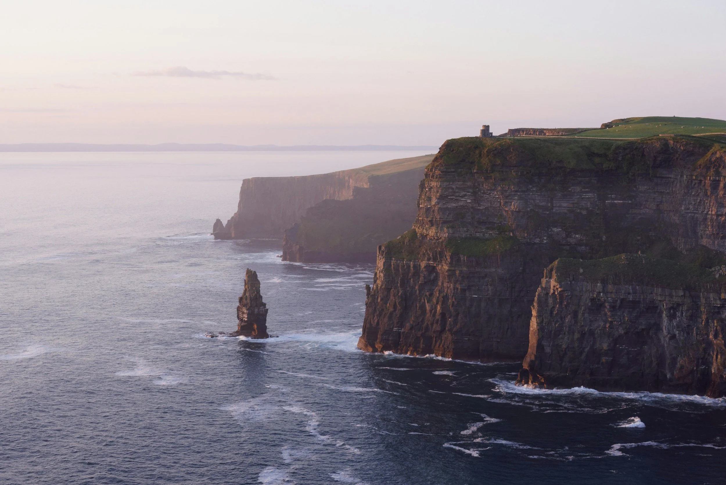 Cliffs along a coastline with the sea below, and a small tower or structure on top of the cliffs, during a time with soft lighting.