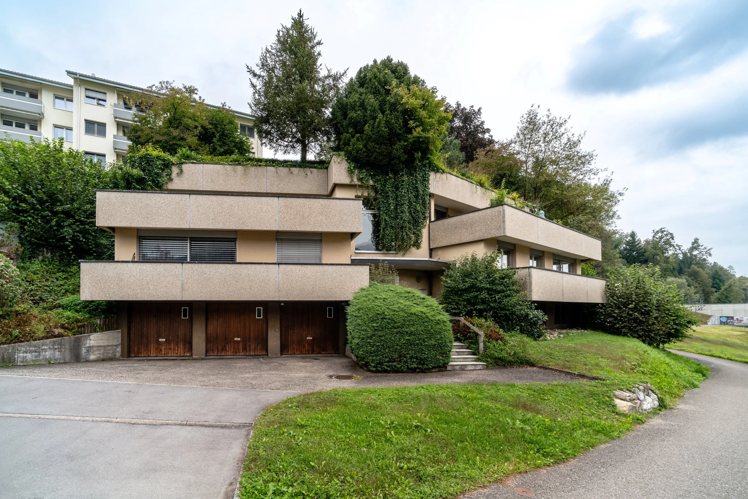 Buchfeldstrasse 23, Buchrain - ZweistöckigesHaus mit großzügigen Terrassen, umgeben von grünen Bäumen und Pflanzen, im Hintergrund ein bewölkter Himmel.