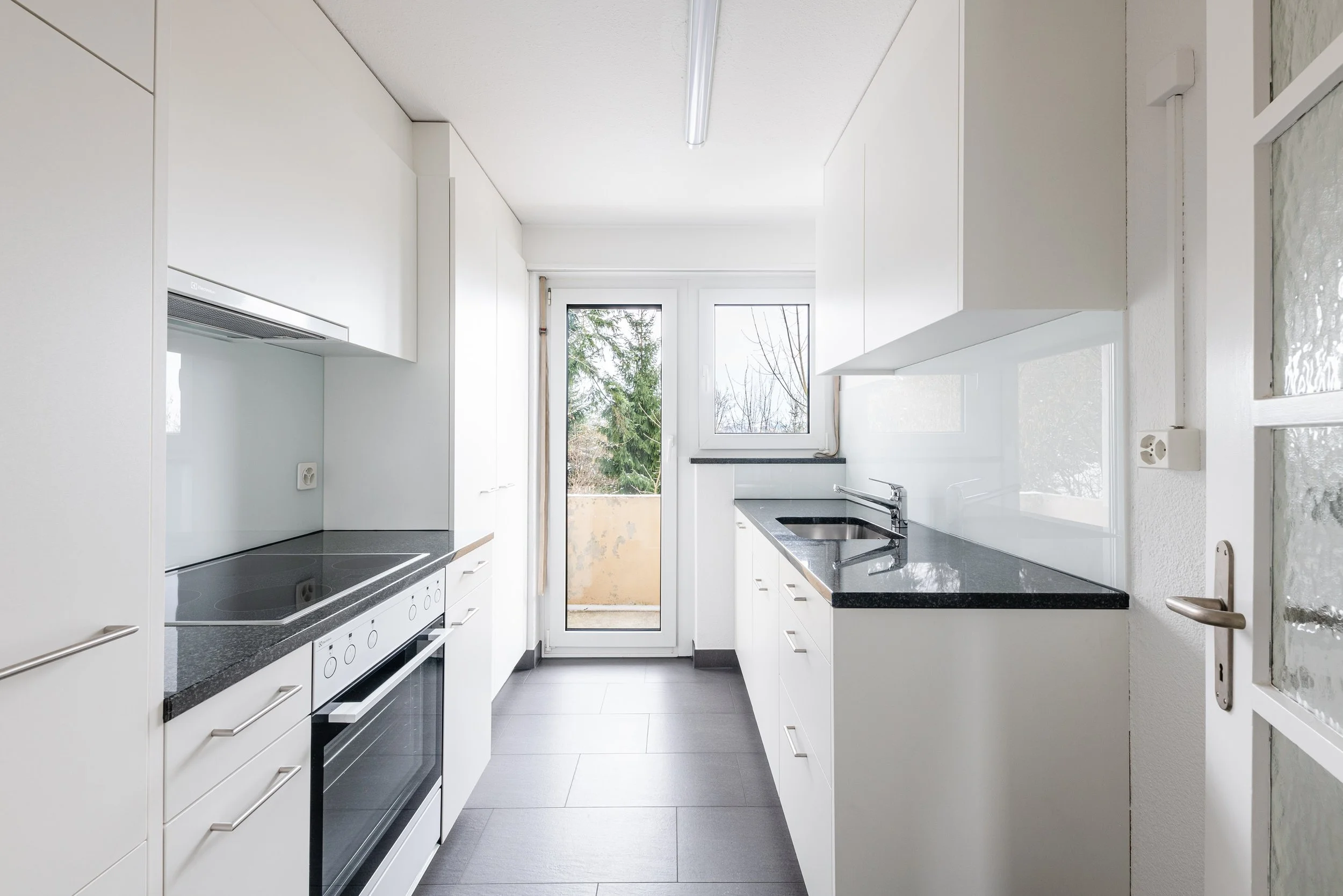 Heiterweid 26/28, Luzern - Küche mit weißen Schränken, schwarzen Arbeitsplatten und einem Fenster, das einen Blick nach draußen ermöglicht.