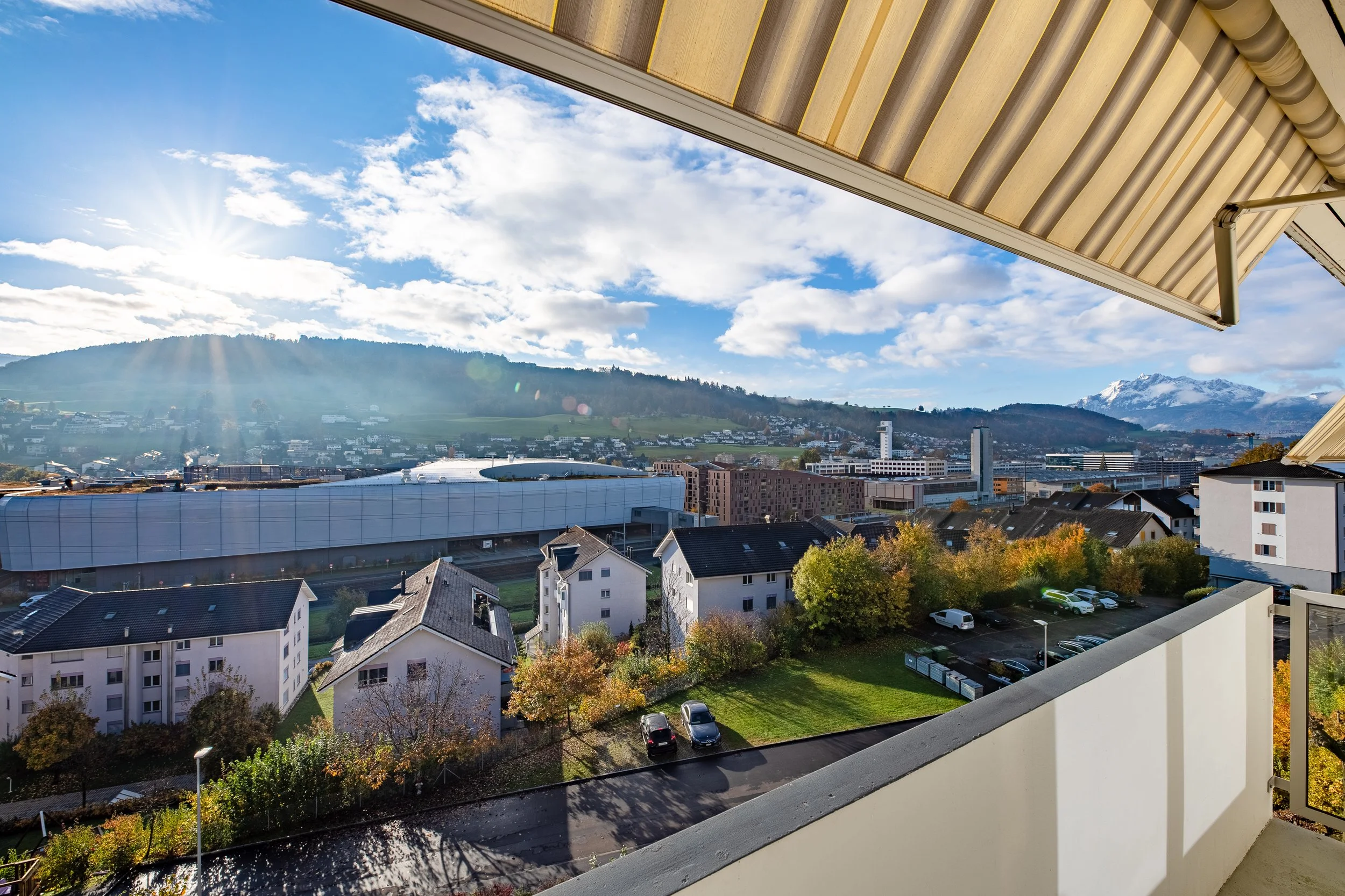 Buchfeldstrasse 15-21, Buchrain - Blick von einem Balkon auf eine Ebikon mit Häusern, Bäumen und großen Bergen im Hintergrund, sonniger Himmel mit Wolken.