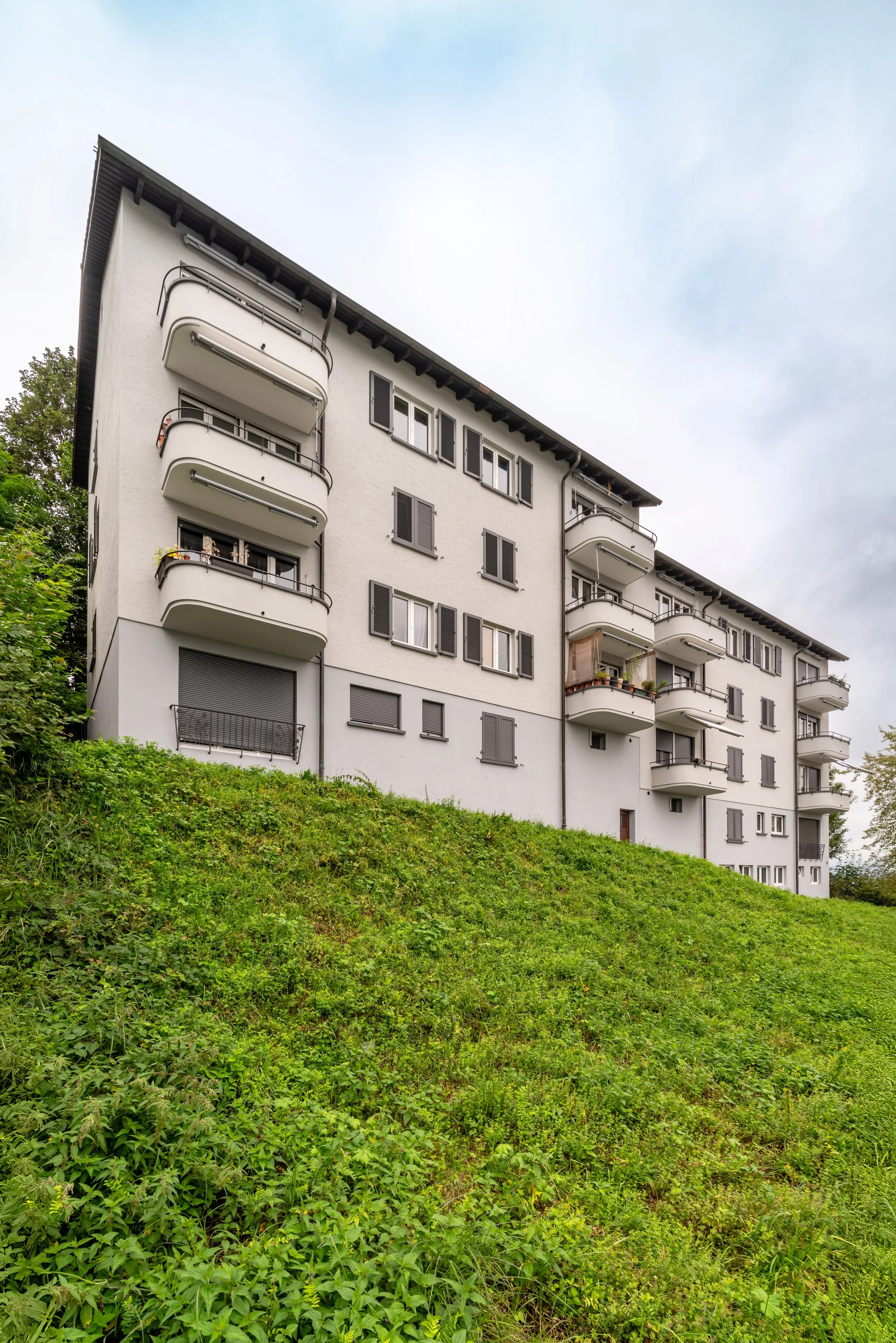 Heiterweid 26/28, Luzern - Mehrstöckiges Wohngebäude mit mehreren Balkonen, umgeben von grünem Hang mit Gras und Pflanzen, unter bewölktem Himmel