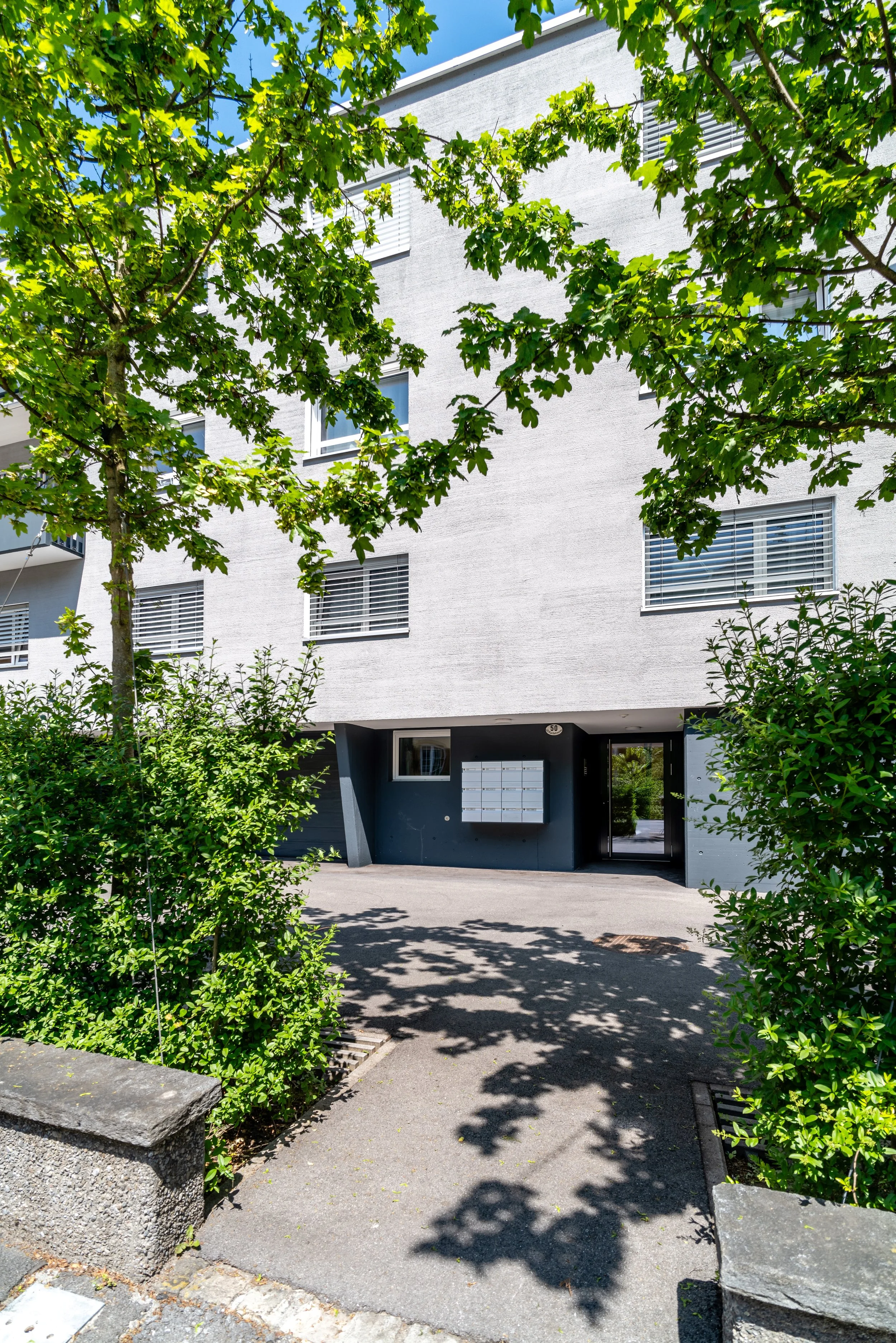 St. Karlistrasse 50, Luzern - Mehrfamilienhaus mit weißen Wänden, mehreren Fenstern mit Rollläden, Eingangsbereich mit Briefkästen, umgeben von grünen Büschen und Bäumen, Schatten auf dem Gehweg bei sonnigem Himmel.