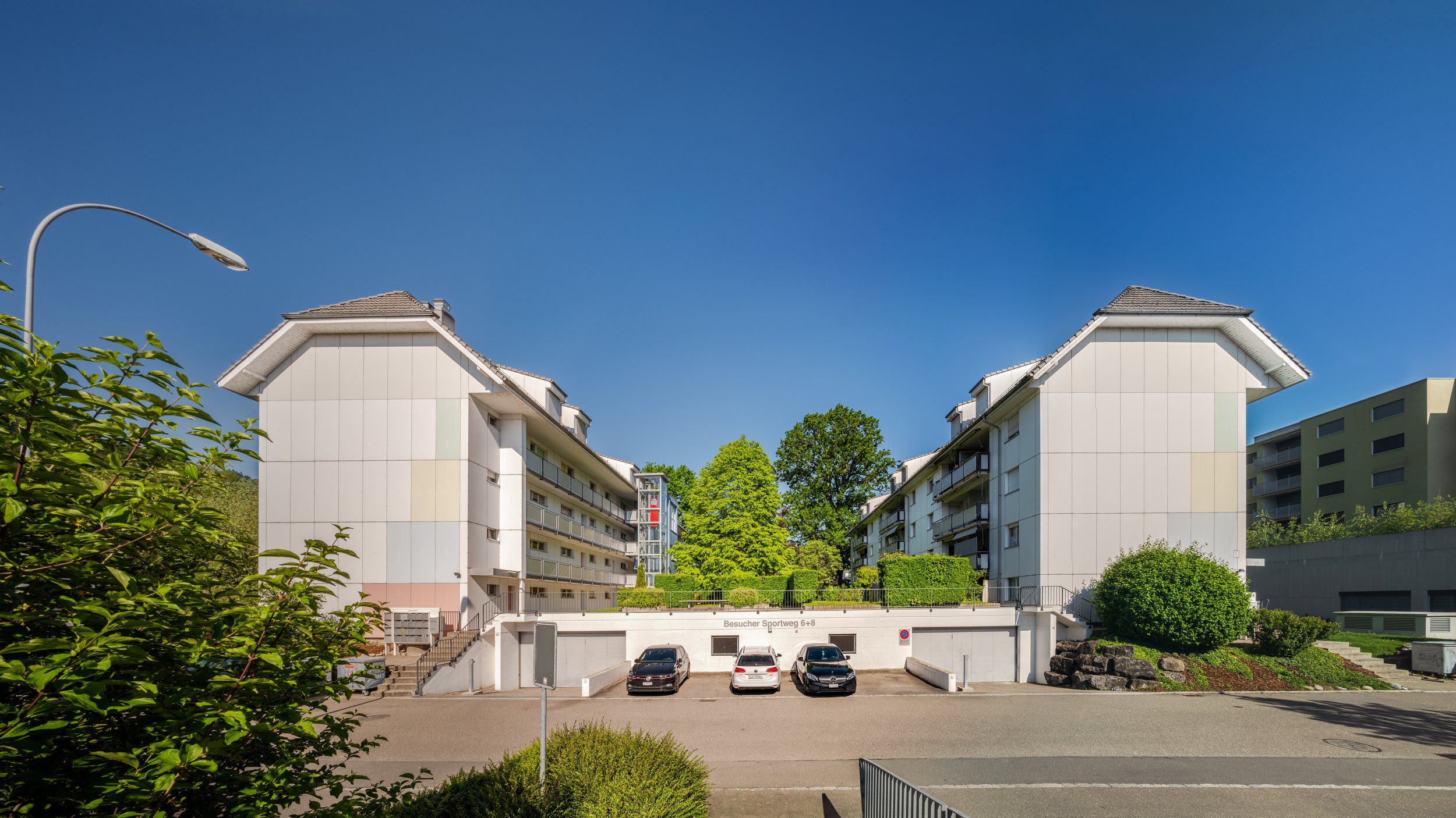 Sportweg 6/8, Kriens - Gerade Ansicht eines Doppelwohnblocks mit weißen Fassaden, Balkonflächen, umgeben von Bäumen und Grünflächen, im Vordergrund ein Parkplatz mit drei Autos.