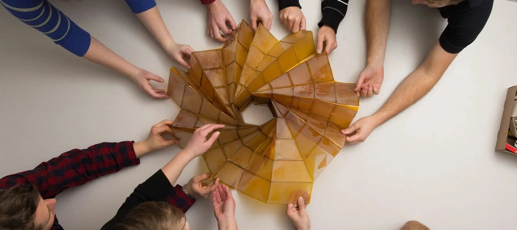 Multiple people holding and assembling a large stained glass mosaic on a white table.