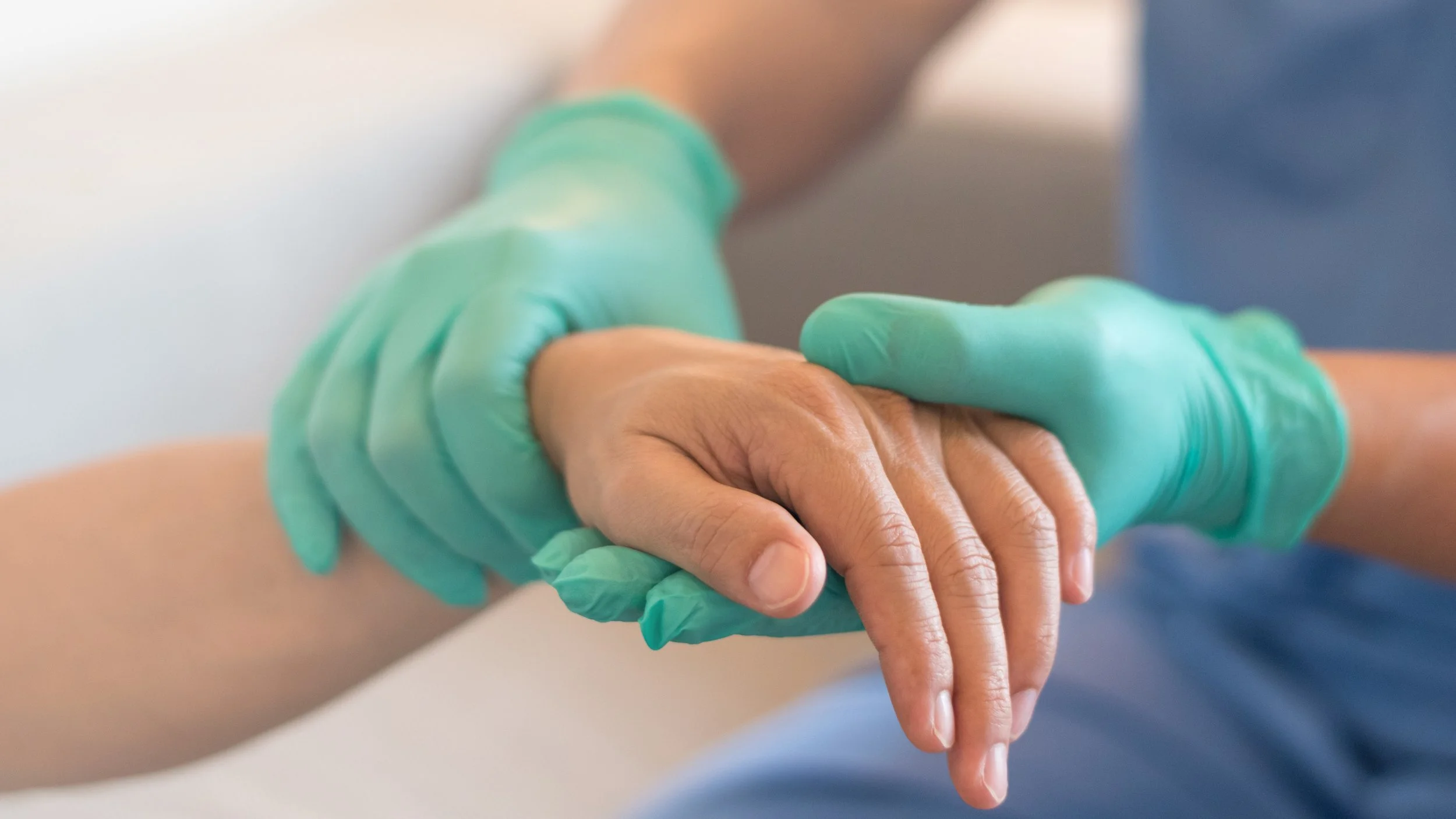 Close-up of a healthcare worker wearing teal gloves gently holding an elderly person's hand.
