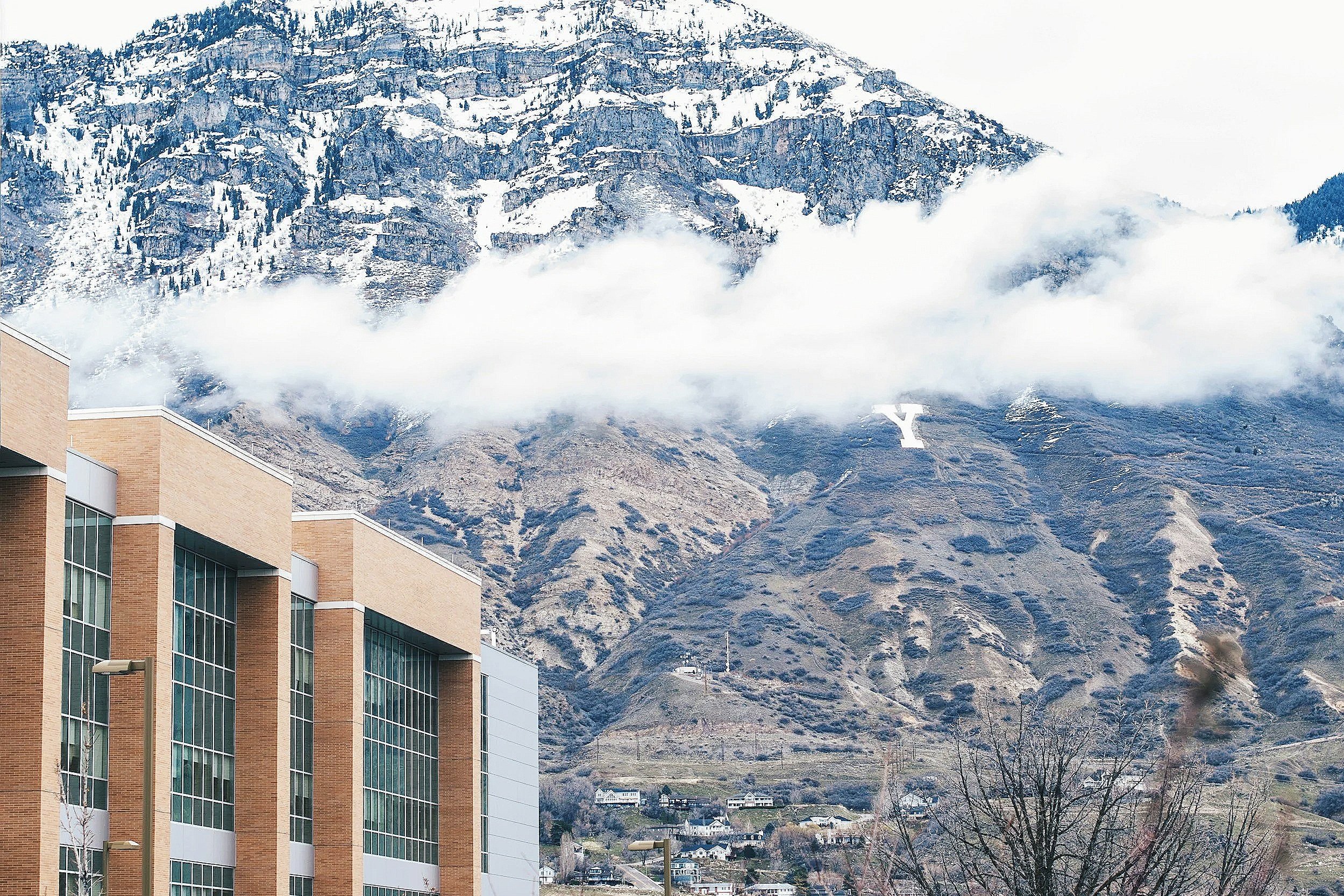 A mountainous landscape with snow-capped peaks, clouds, and a large 'Y' sign on the hillside. In the foreground, there are modern buildings with beige brick facades and large glass windows.