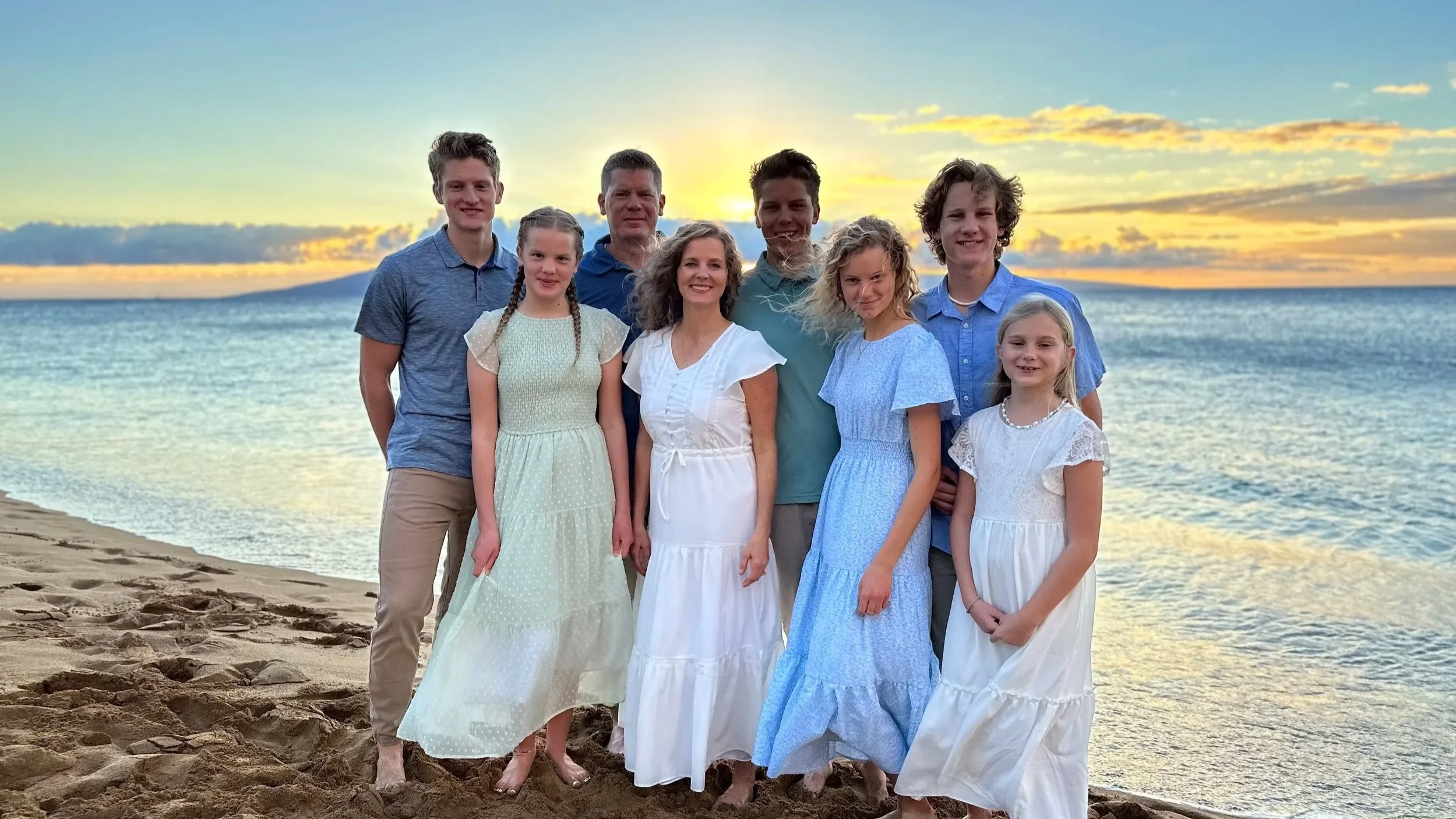 Group of nine people, including children and adults, standing on a sandy beach at sunset with the ocean and cloudy sky in the background.