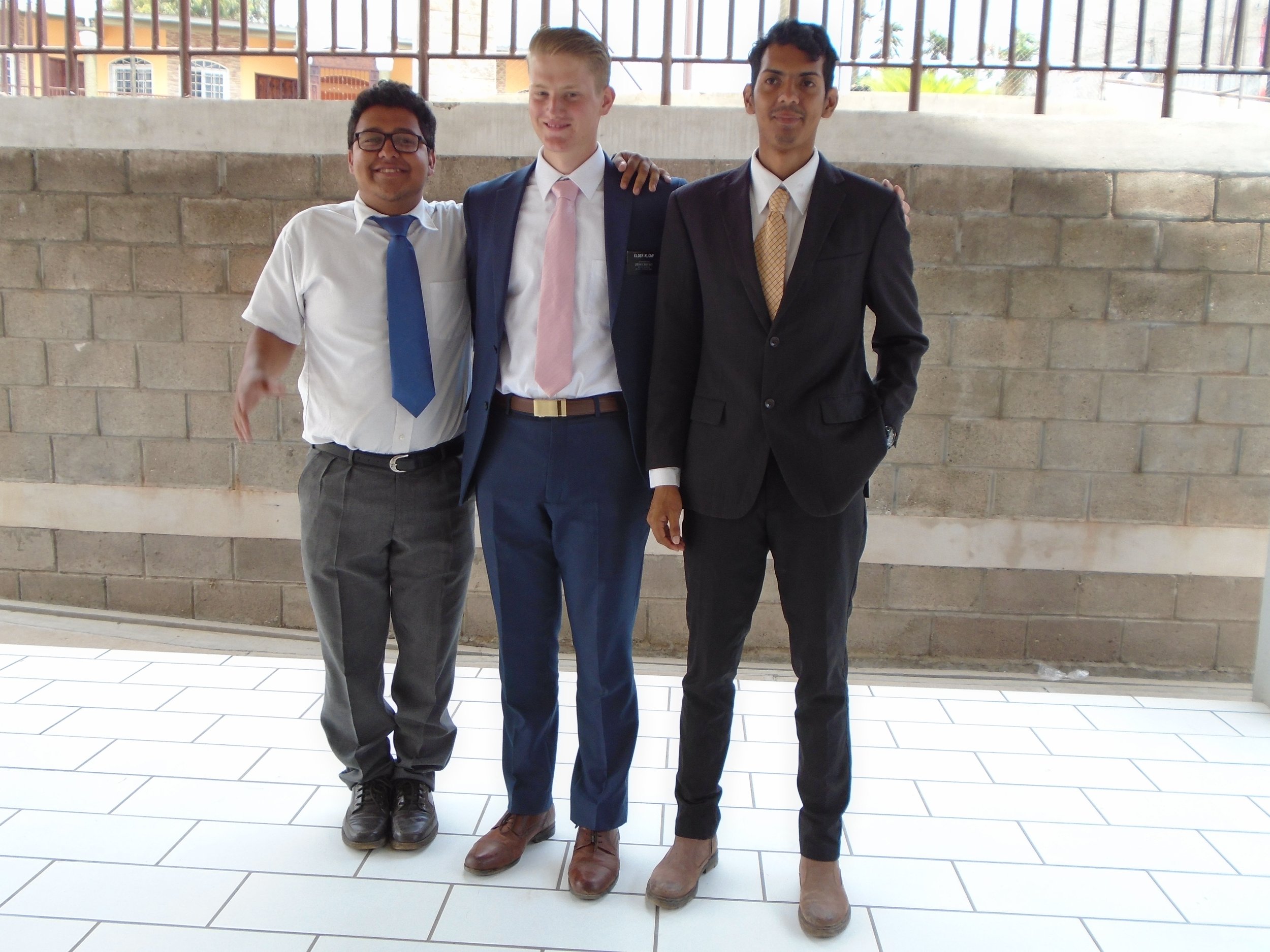 Three young men dressed in formal attire standing together outdoors in front of a brick wall and metal railing, with a building visible in the background.