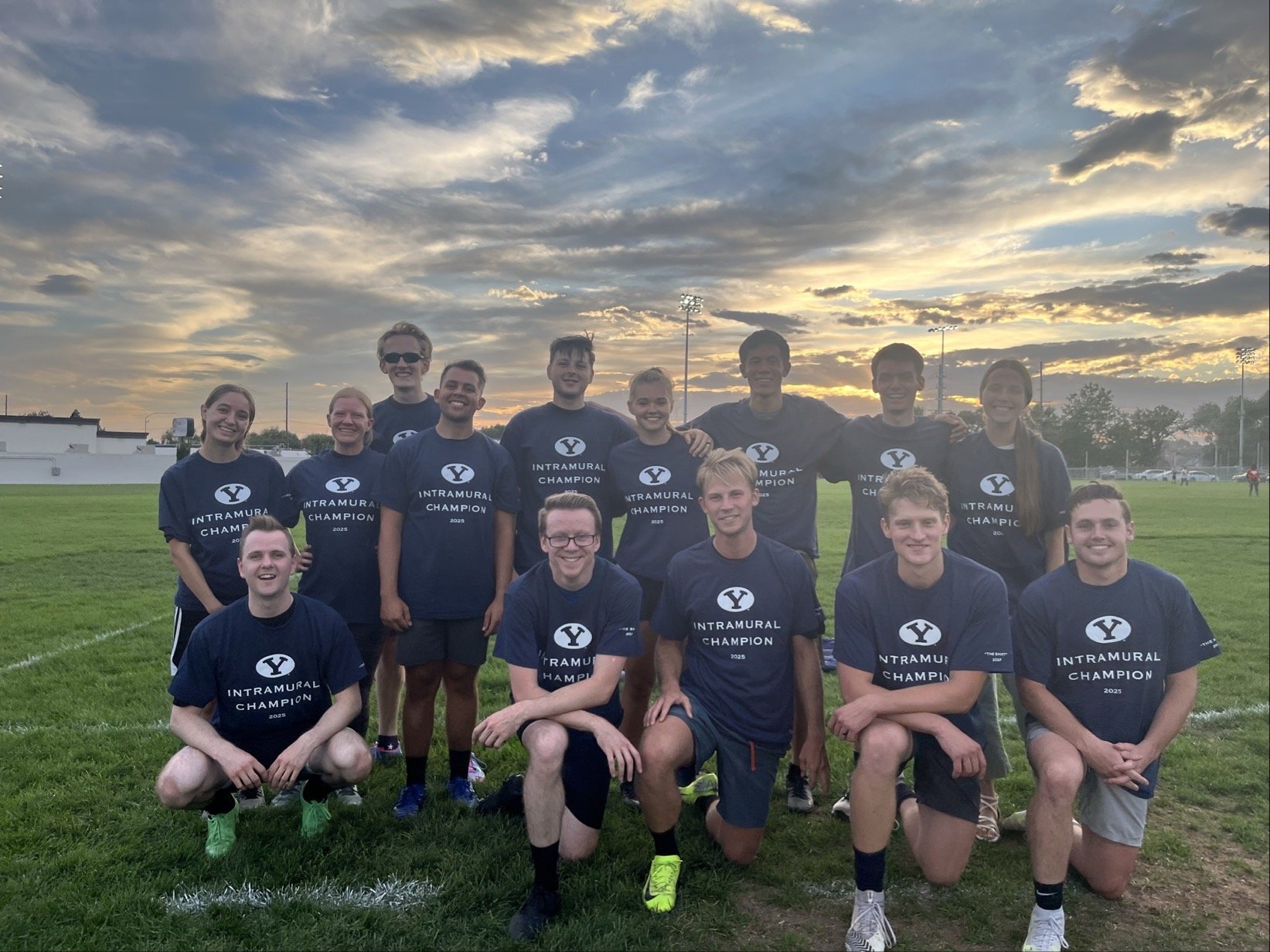 Group of young athletes on a grassy field at sunset, wearing the BYU intramural "The Shirt"