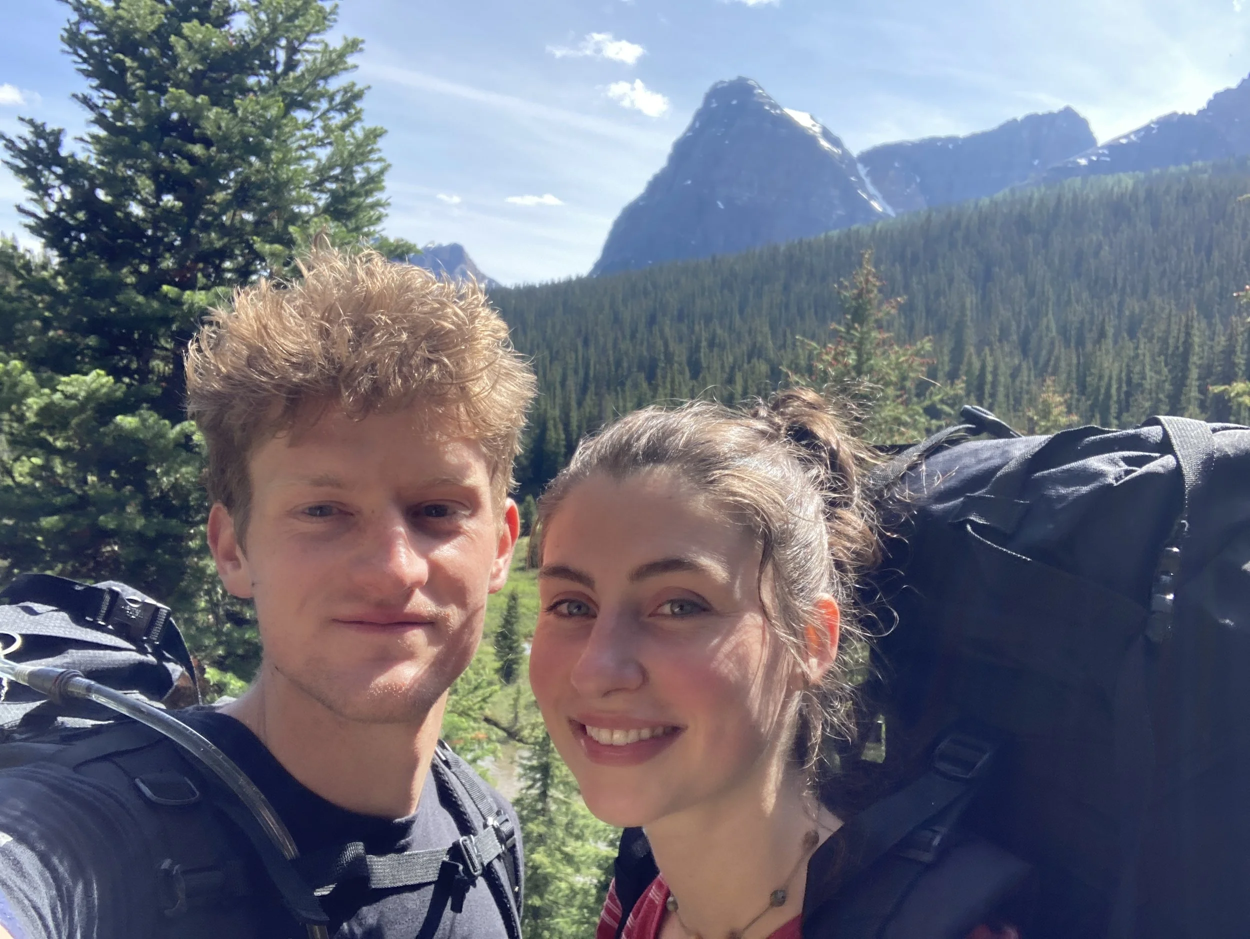 Two young hikers with backpacks smiling outdoors in a forested mountain landscape with tall peaks and greenery.