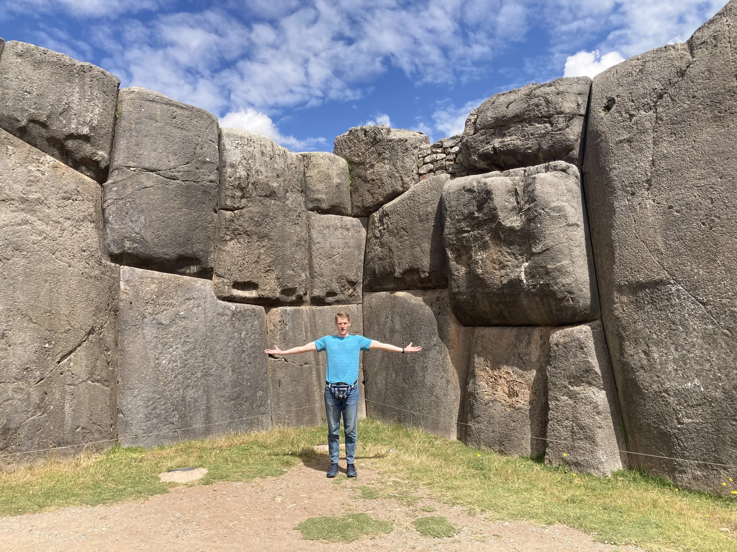 A person in a blue shirt and jeans stands with arms outstretched in front of ancient stone megaliths under a partly cloudy sky.