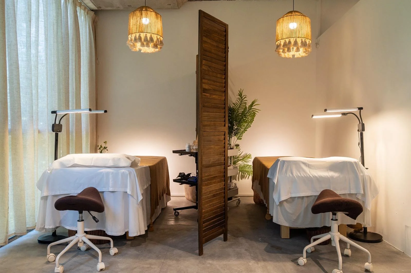 A room with two massage tables separated by a wooden room divider, each table covered with white sheets and brown covers, with adjustable lamps and stools beside them. There are potted plants and large curtains on the window.