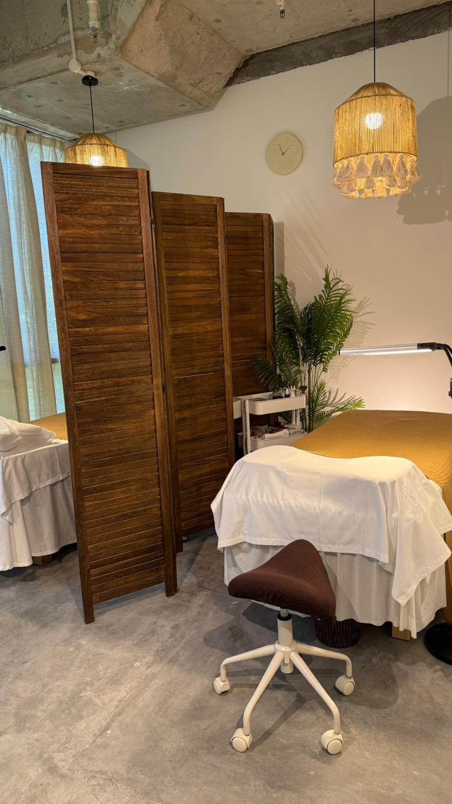 Interior of a room with a massage table covered with white and yellow sheets, a brown rolling chair, a wooden room divider, a white clock on the wall, a potted plant, and two hanging lamps with woven shades.