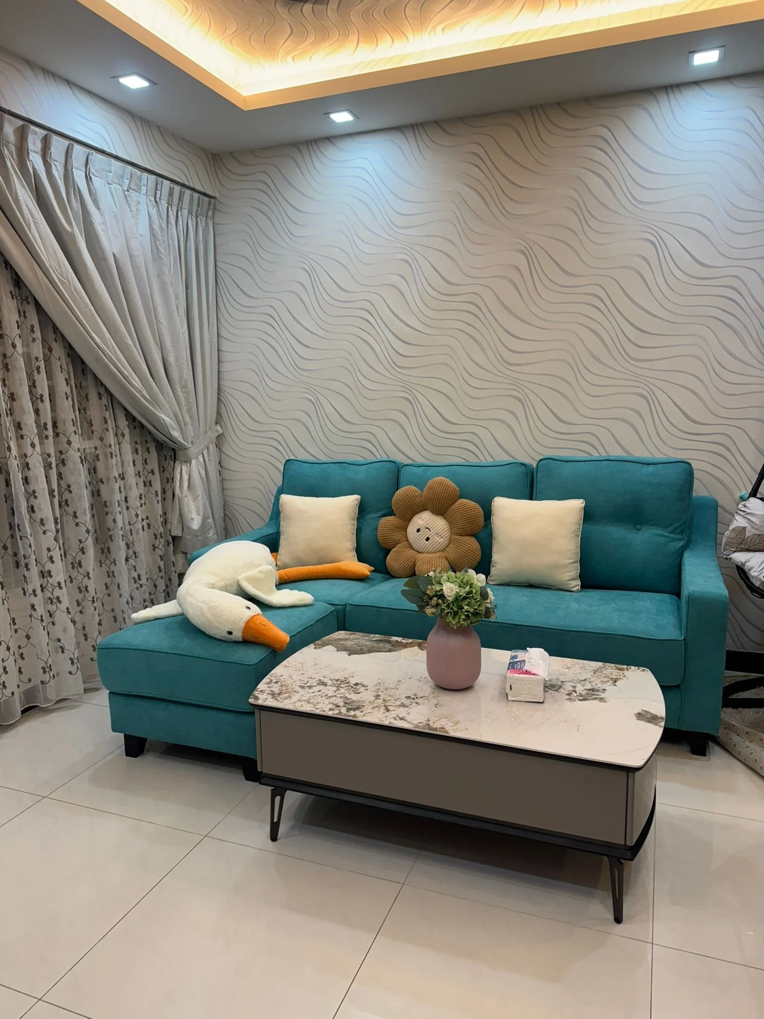 Living room with a turquoise sofa decorated with plush toys and pillows, a marble-top coffee table with a pink vase of flowers, cream curtains, and a textured wall with warm ceiling lighting.