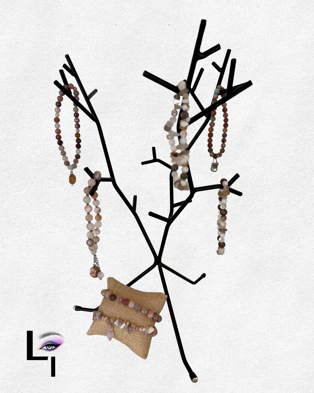 Black jewelry stand shaped like a tree displaying various beaded necklaces and bracelets, with a small burlap pillow holding additional jewelry, set against a plain white background.