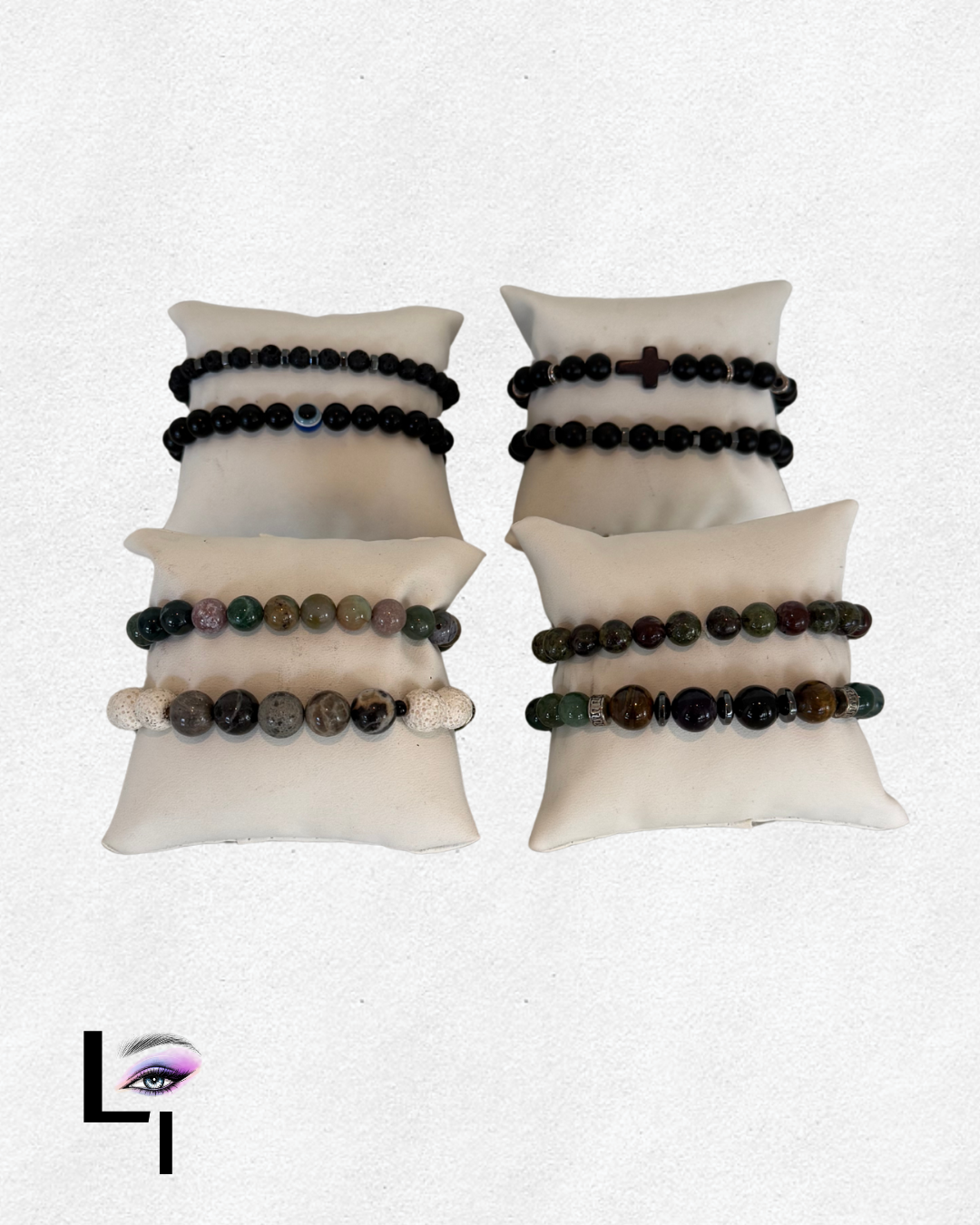 Four bead bracelets displayed on white pillow cushions, with a logo of an eye within an 'L' in the bottom left corner. The bracelets feature various styles and colors of beads, some with additional charms.