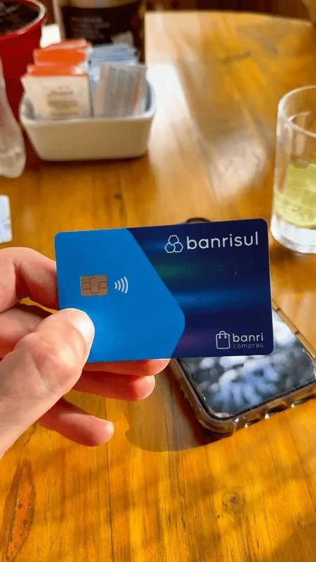 Hand holding a blue Banrisul debit card with a contactless symbol, on a wooden table with a smartphone, a glass of light-colored beverage, and a container of condiment packets.