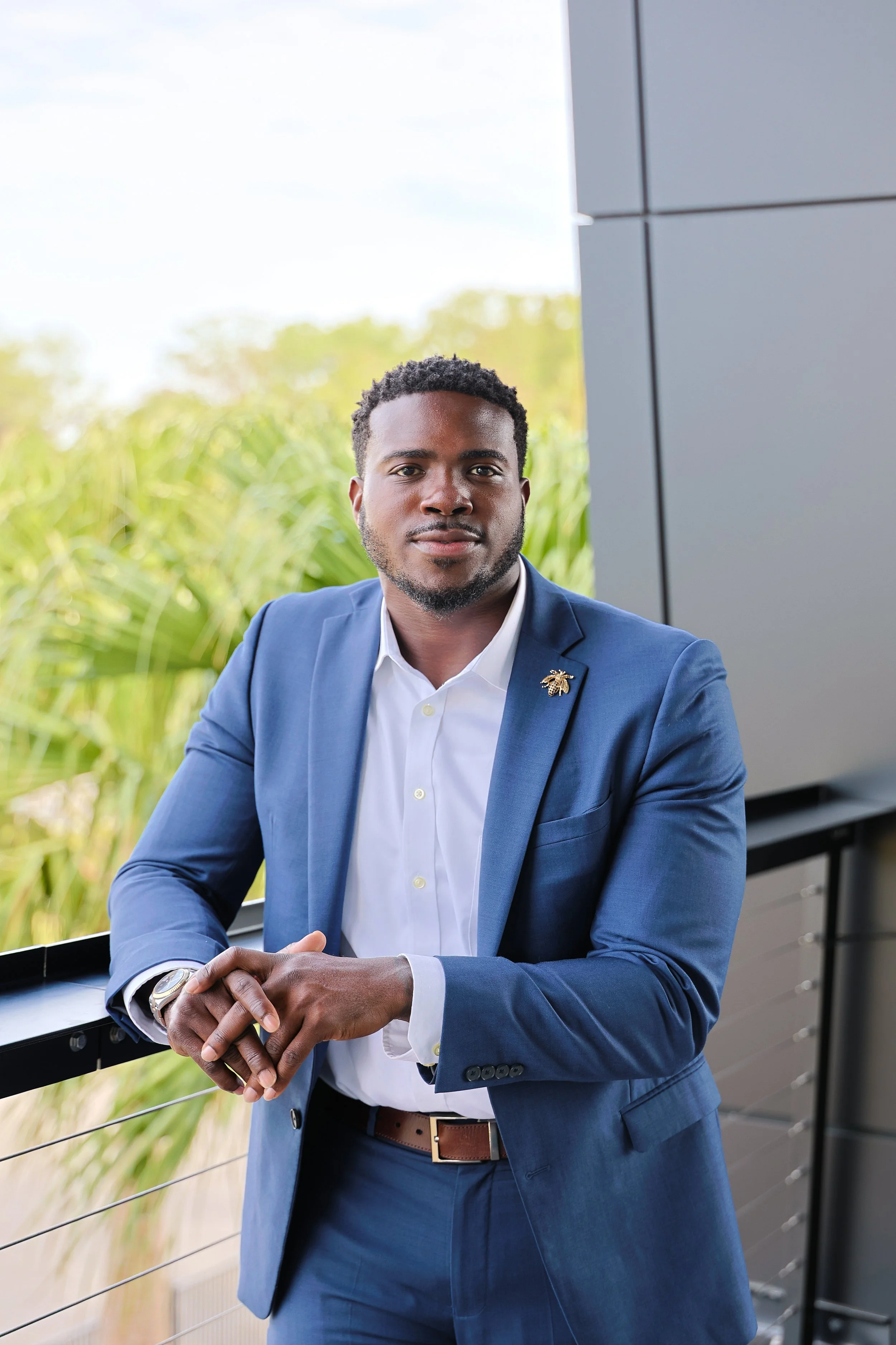 A man in a blue suit sitting outdoors, looking at the camera with a neutral expression. He is dressed professionally, wearing a white shirt and a watch on his left wrist, with a background of greenery and a modern outdoor setting.