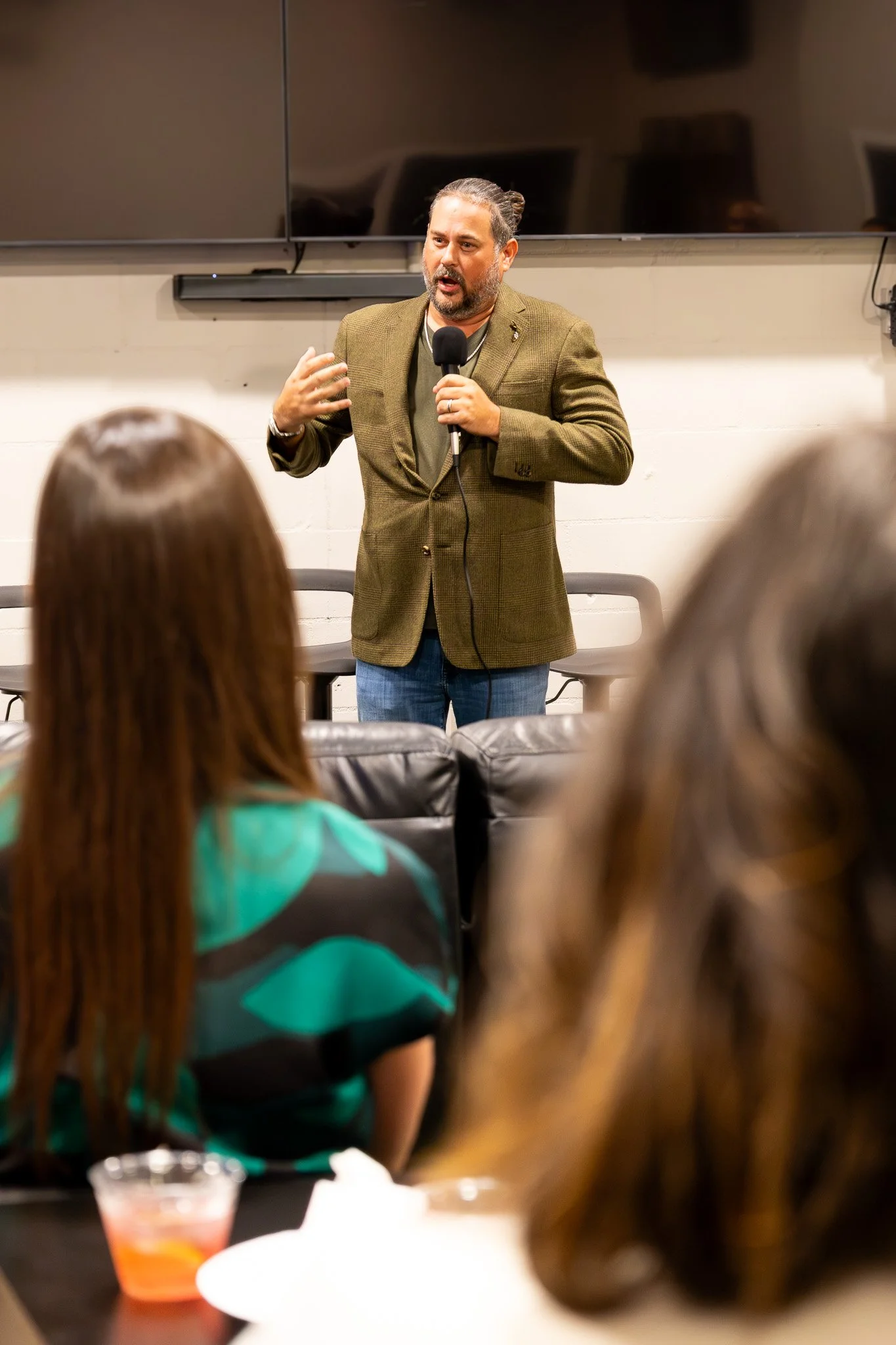A man with a beard and long hair in a bun is speaking into a microphone in front of an audience. There is a black flat-screen TV mounted on the wall behind him, and the focus is on the speaker.