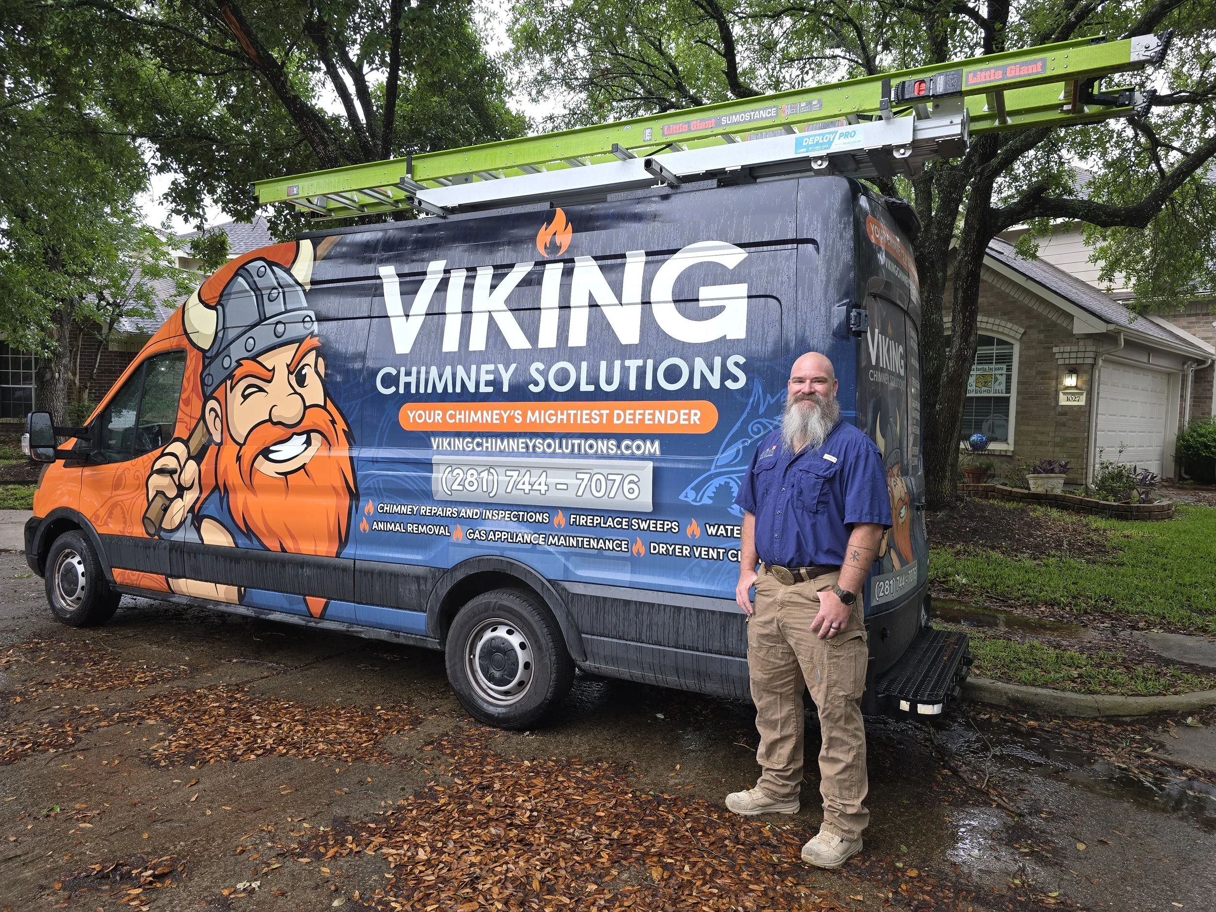 A man with a beard and tattooed arm standing next to a branded service truck for Viking Chimney Solutions, which has a graphic of a Viking with a helmet and axe, parked in front of a residential home with trees and house in background.
