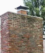 Brick chimney on a house with a black metal chimney cap, surrounded by green trees.