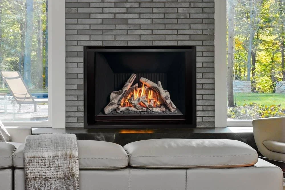 Living room with a fireplace and logs, white sofa, and large windows showing trees outside.