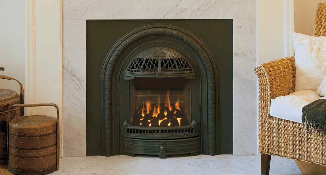 Fire burning in a decorative indoor fireplace with a black metal mantel, surrounded by cream-colored walls and marble accents, with wicker furniture nearby. Valor Fireplace