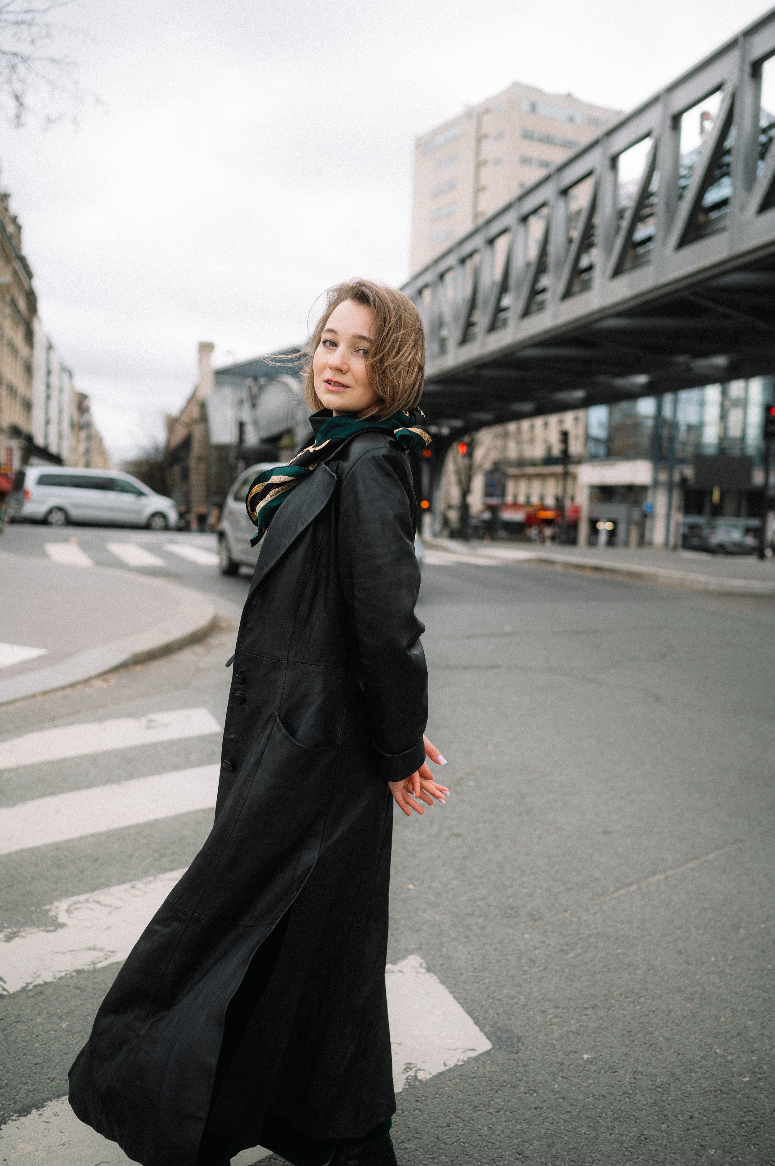 A young woman with shoulder-length hair stands on a city street crossing, wearing a long black coat and a scarf, with buildings and a bridge overhead in the background.