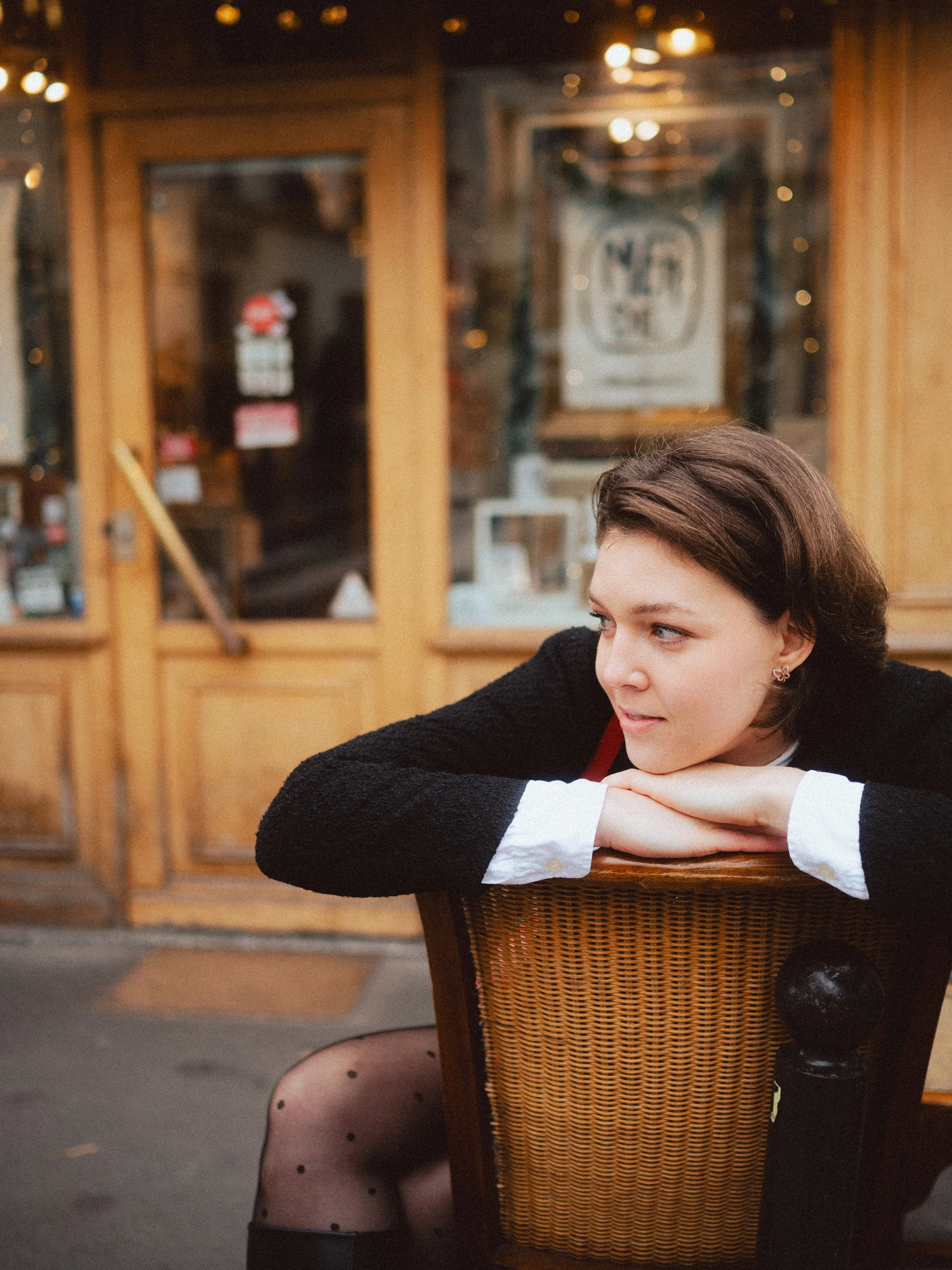 A woman with short brown hair, wearing a black cardigan and white shirt, is resting her chin on her folded arms on the back of a chair outside a wooden storefront, looking to her right.