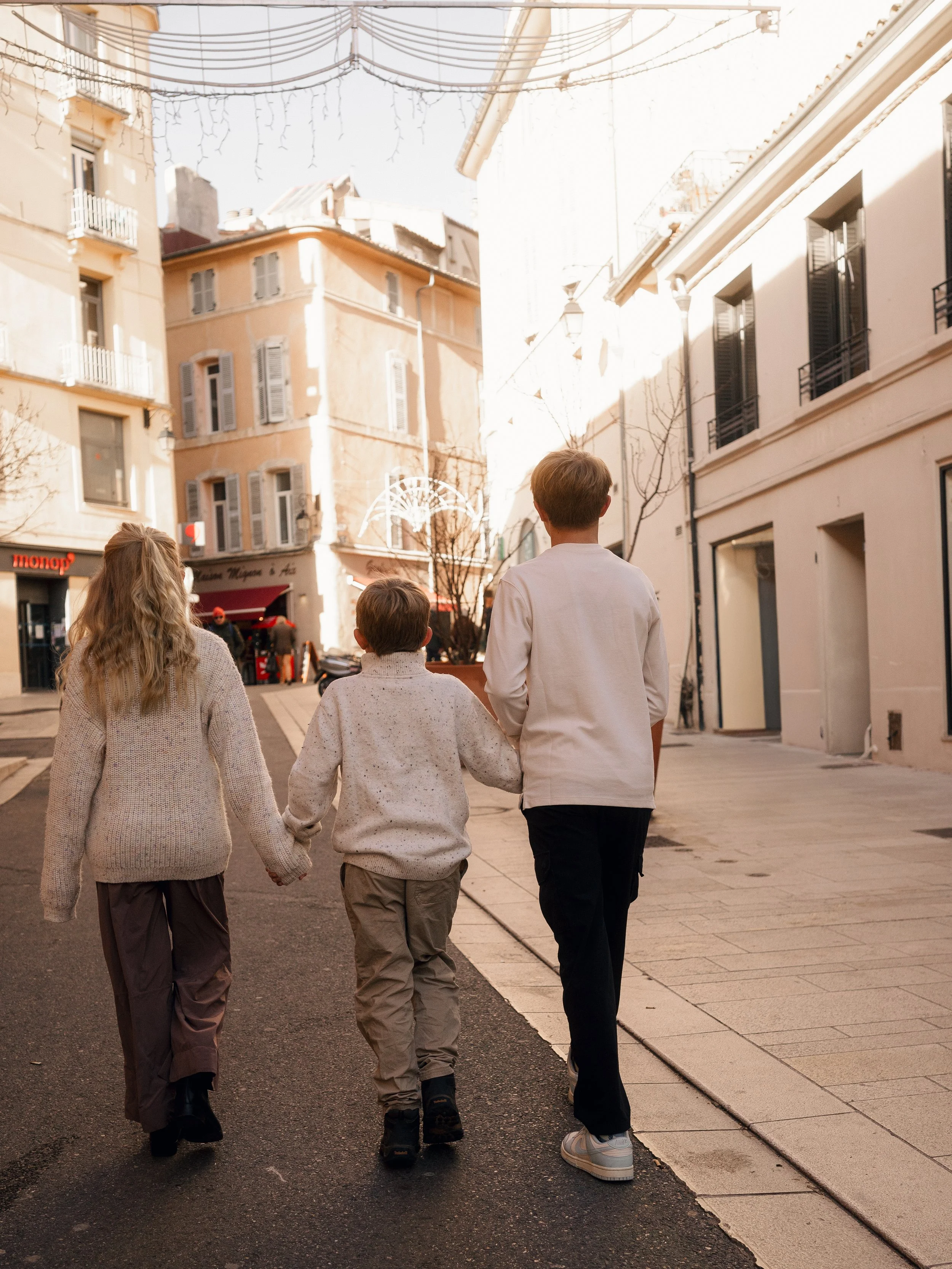 Three children walking together down a city street on a sunny day, holding hands.