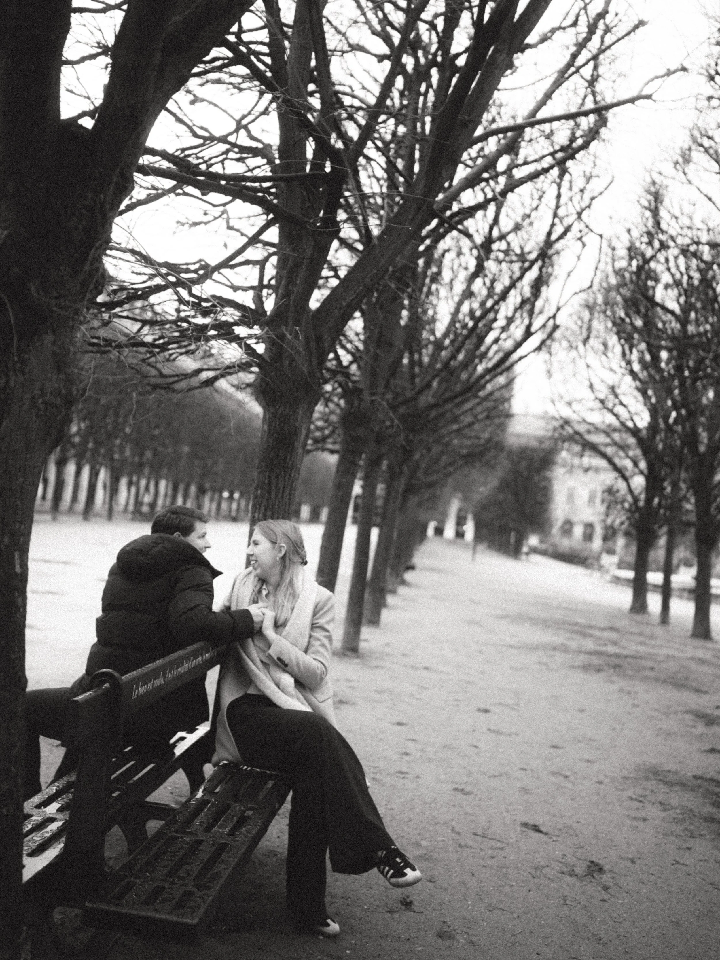 A black and white photo of a man and woman sitting on a park bench, smiling and holding hands, under leafless trees on a cloudy day.