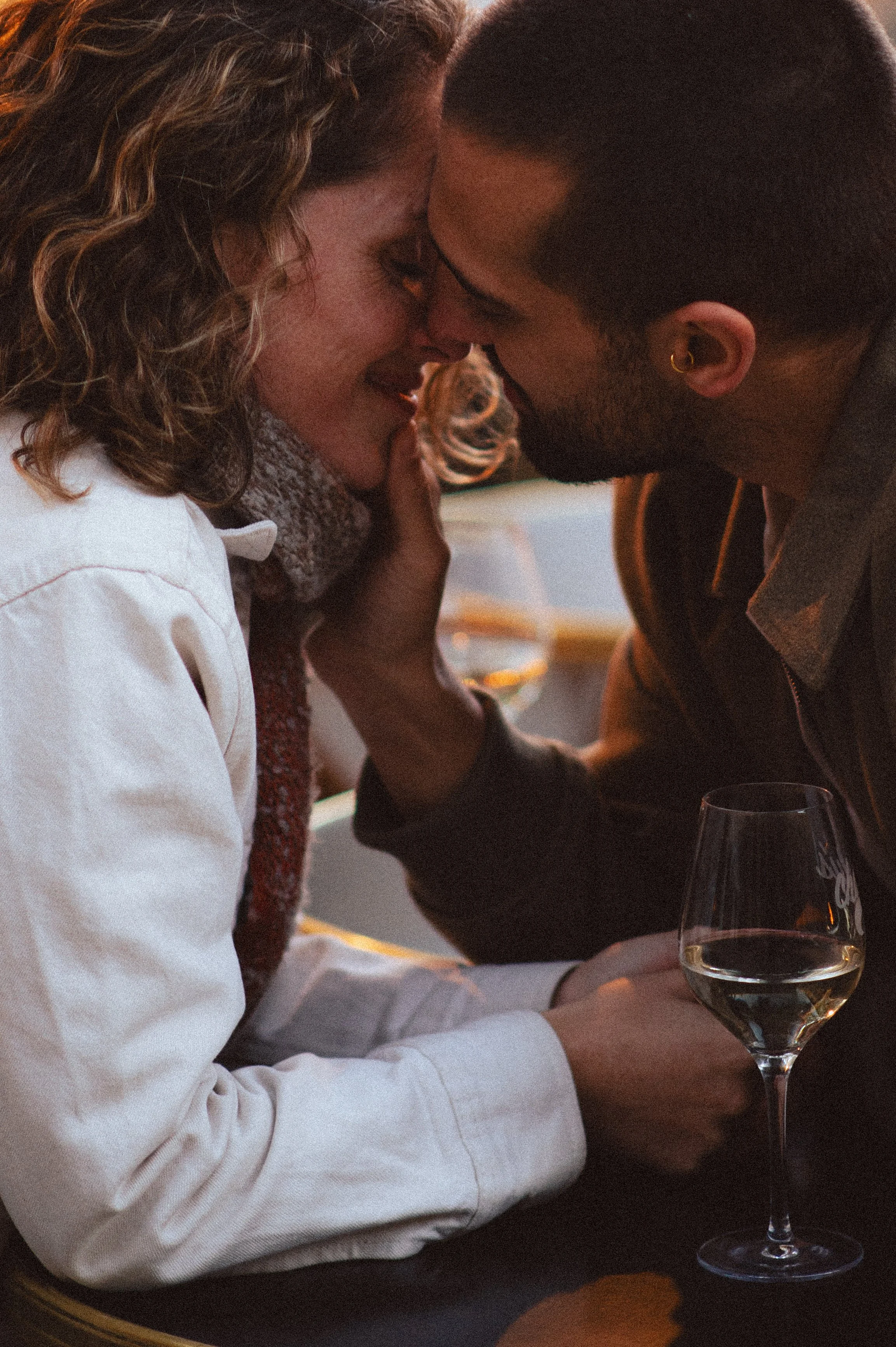 A couple sharing an intimate moment, with faces close and eyes closed, in a warm ambiance, holding a glass of white wine.
