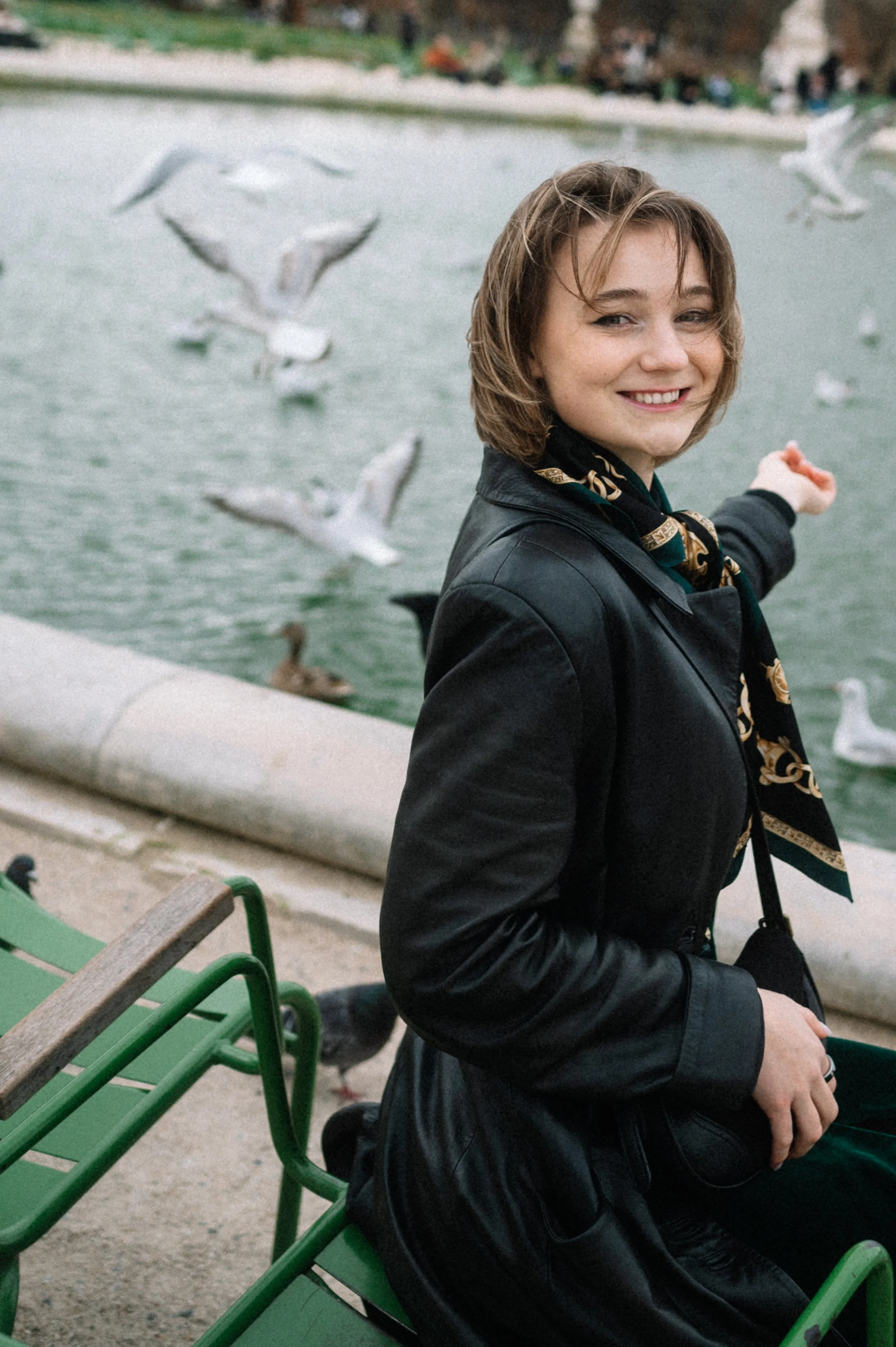 A woman sitting on a green park bench by a lake, smiling and pointing toward a flock of seagulls flying nearby.