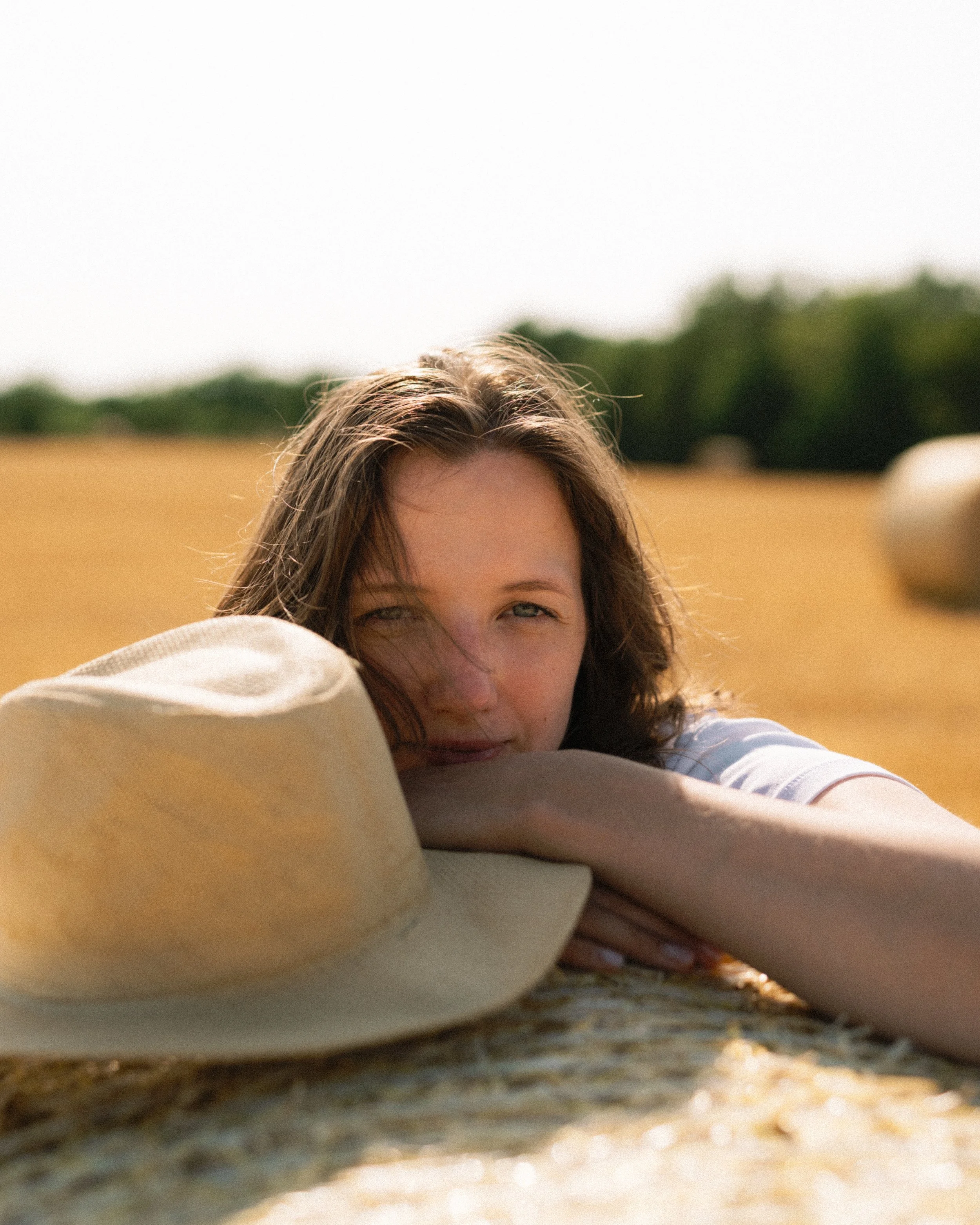 A young woman with brown hair resting her head on a straw hat lying on a hayfield, with a blurred background of trees and a bright sky.