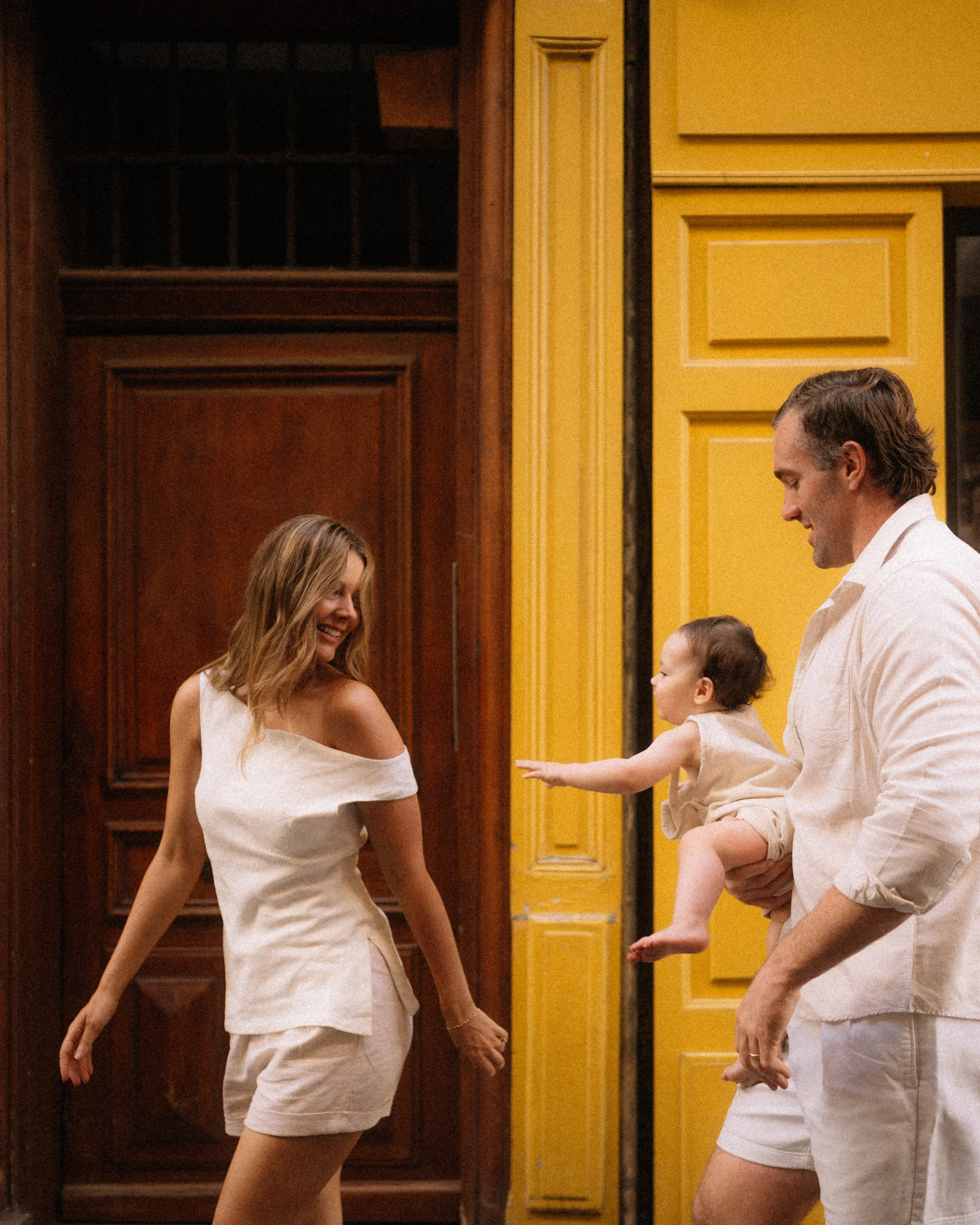 A woman, a man, and a young child are enjoying a happy moment indoors, with the woman smiling and the man holding the child as the child reaches out toward the woman.