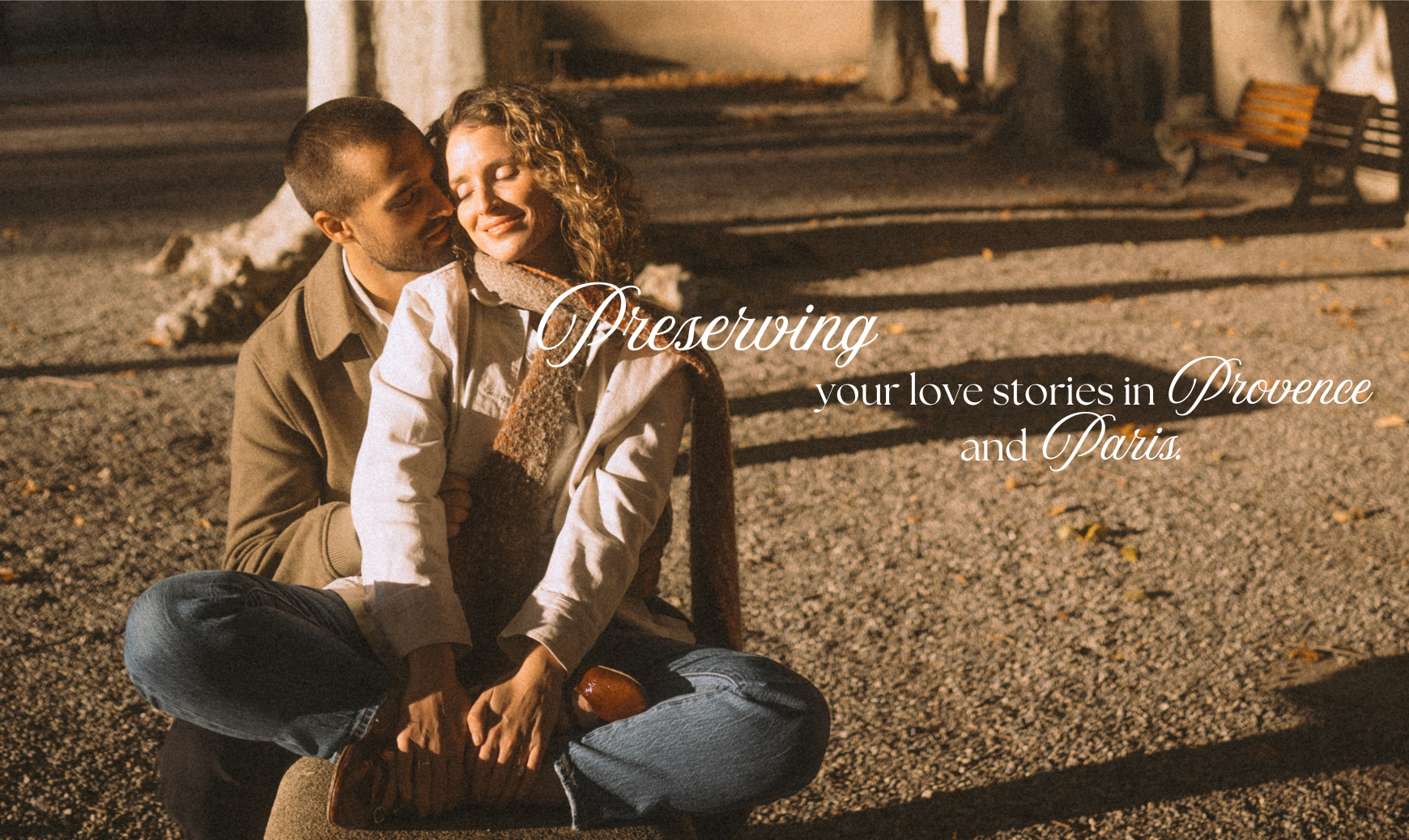 A couple sitting close together on a park bench at night, embracing and smiling, surrounded by autumn leaves and trees.