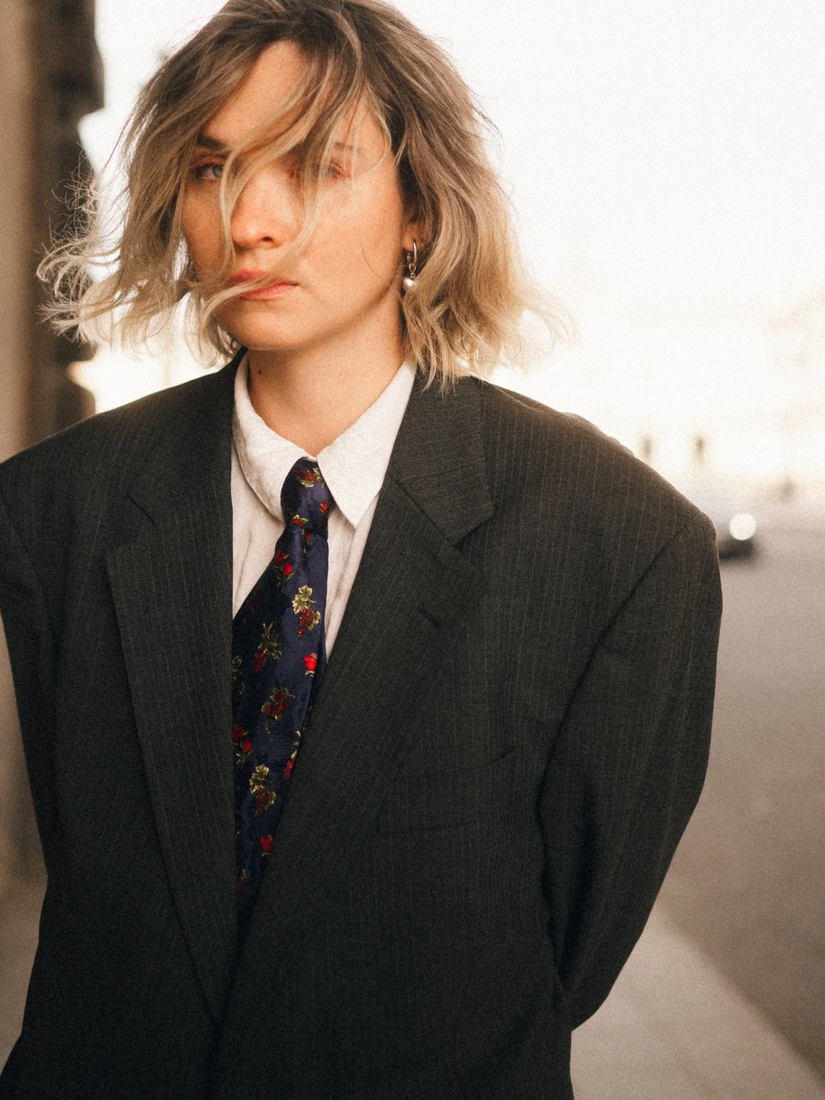 A woman with blonde, wavy hair over one eye, wearing a black blazer, white shirt, and a floral tie, standing outdoors on a city street at sunset.