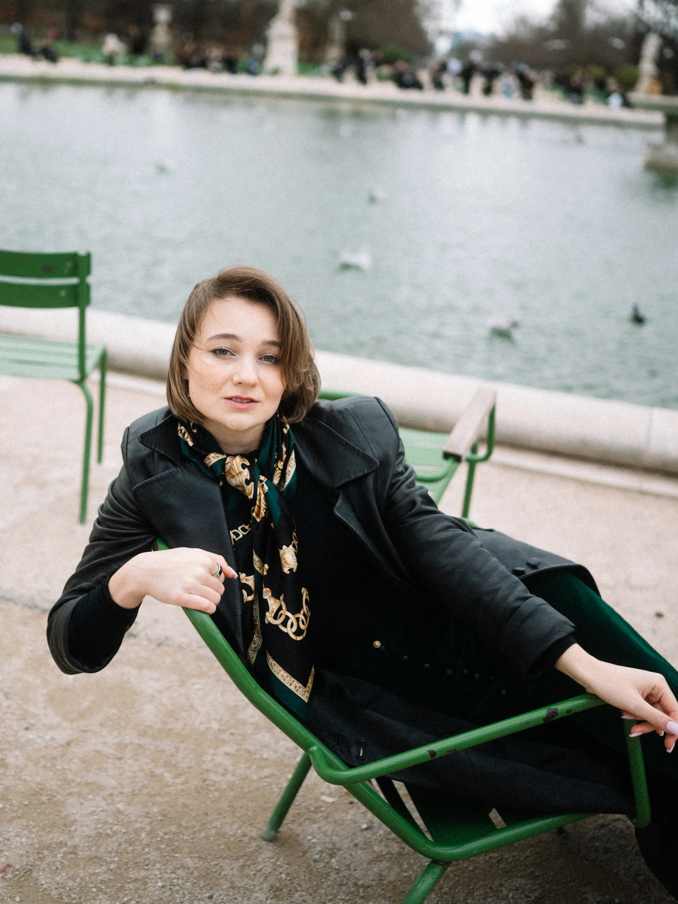 A young woman with short brown hair lounging on a green park bench near a body of water with ducks, in an outdoor park setting.