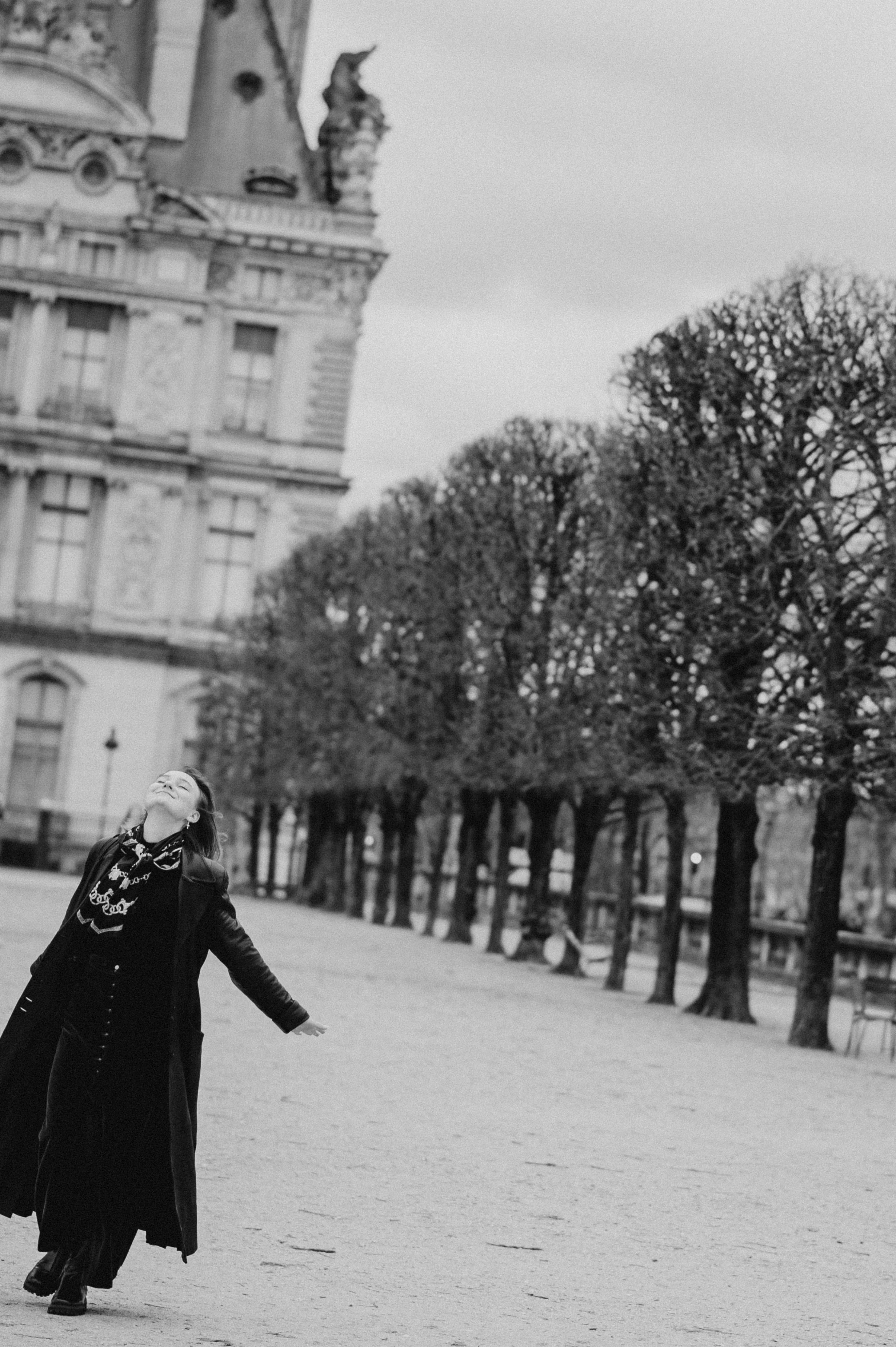 Woman enjoying outdoors, standing with arms open, in front of trees and historic building in black and white.