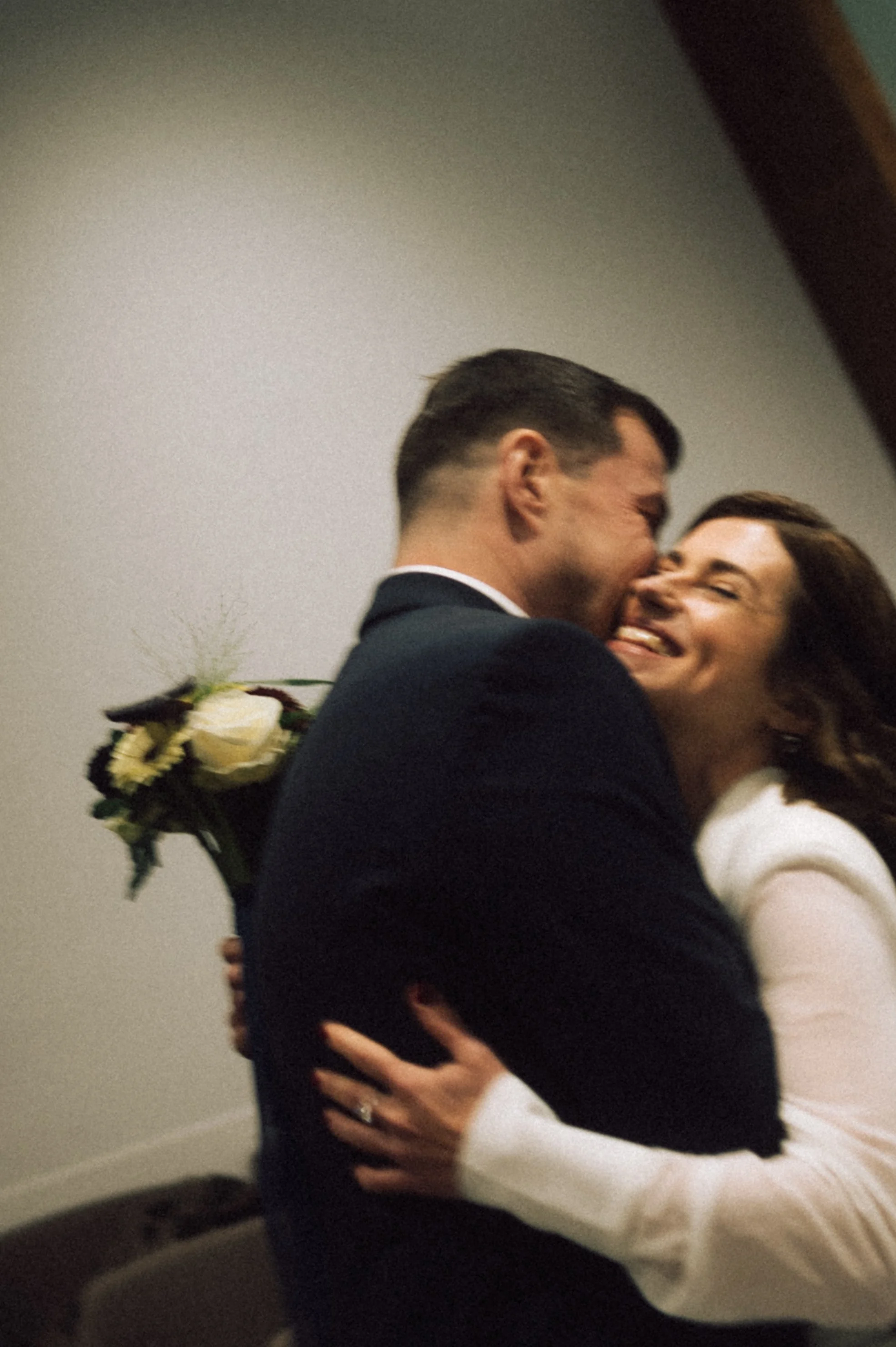 A joyful couple hugging tightly, smiling, with the man holding a bouquet of white roses behind his back.