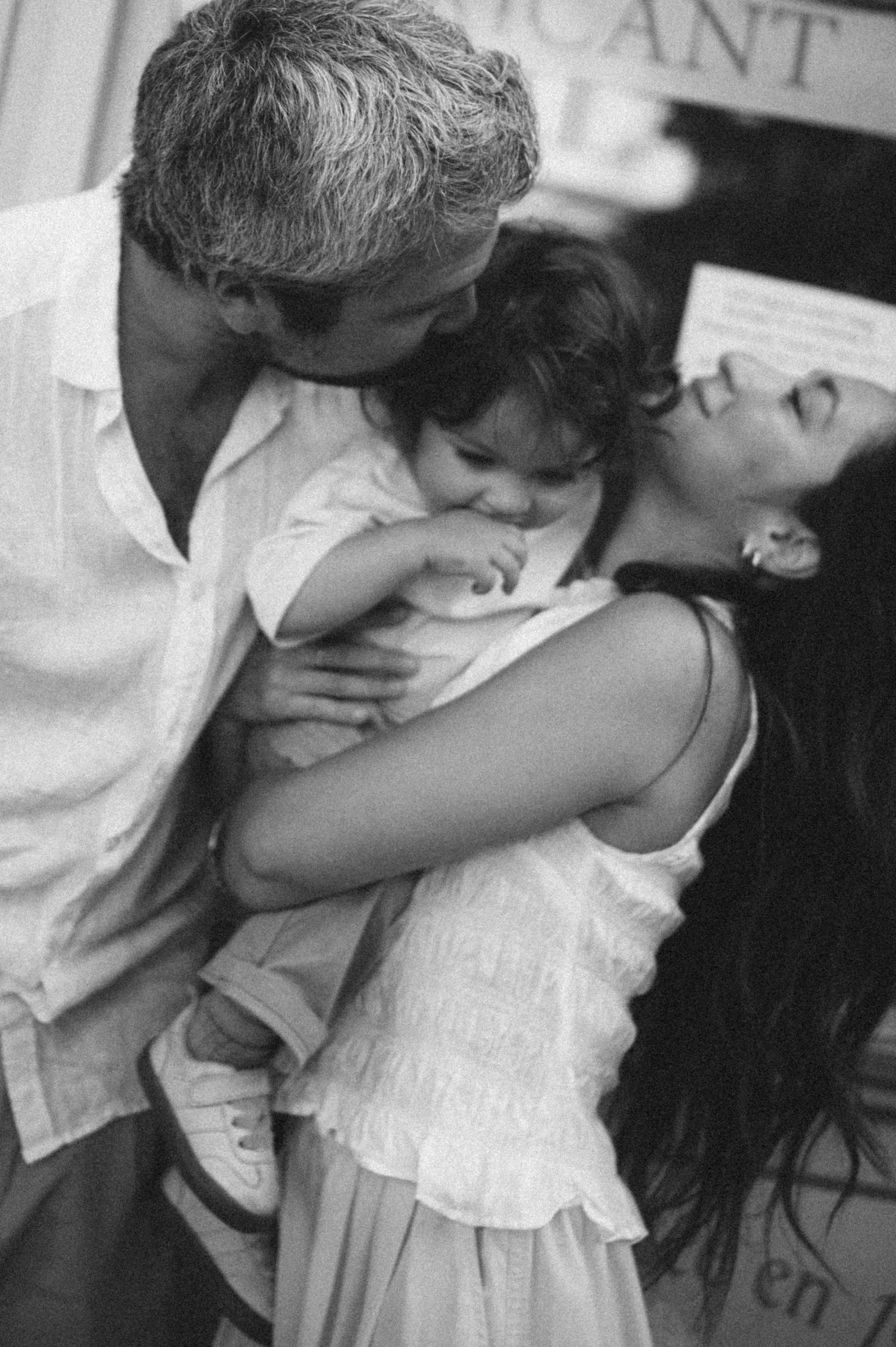 A man, woman, and child in an embrace, with the man kissing the child who is lying on the woman's chest, in a close and affectionate moment.