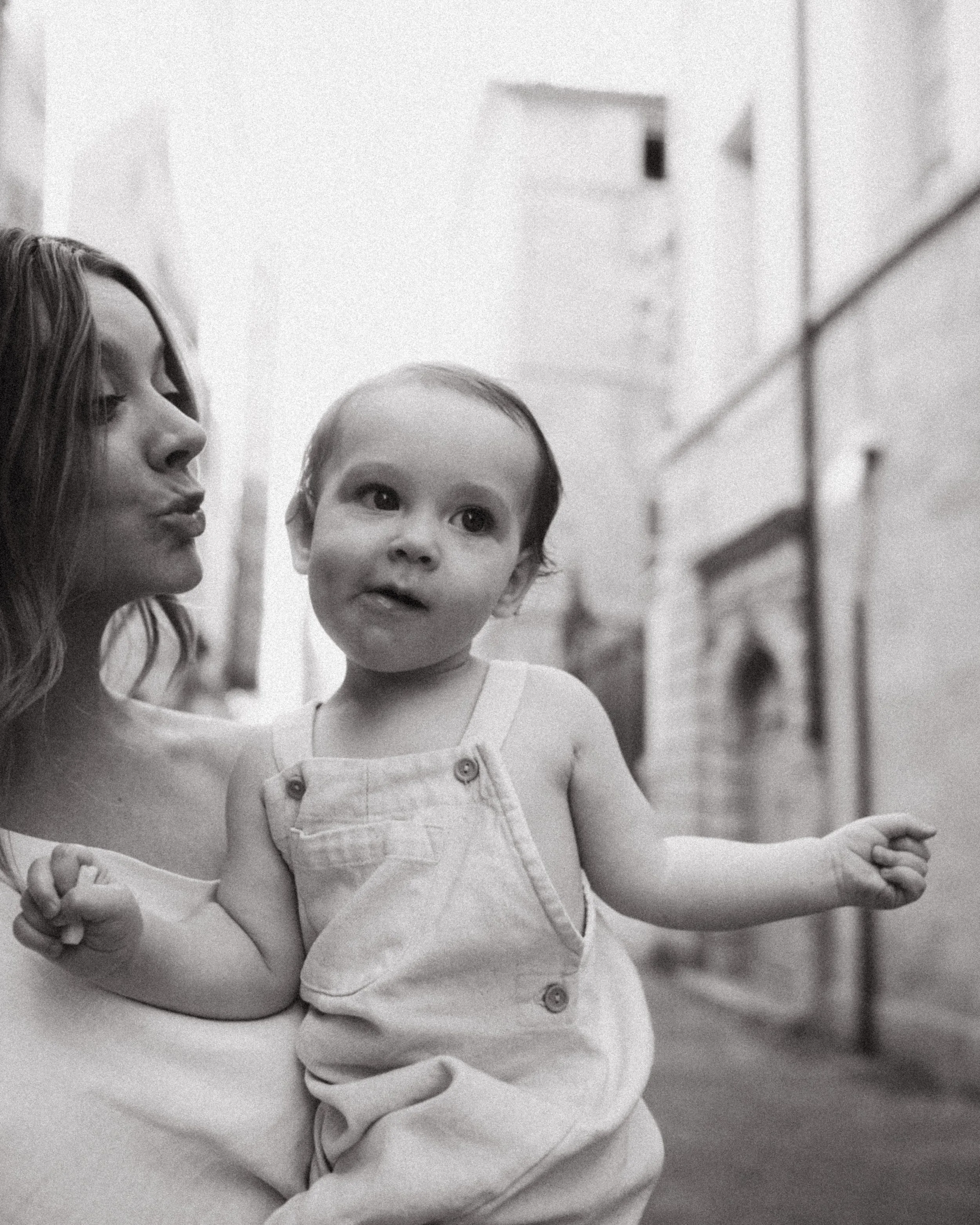 A woman holding a young girl outdoors in an alleyway, both making playful faces at the camera.