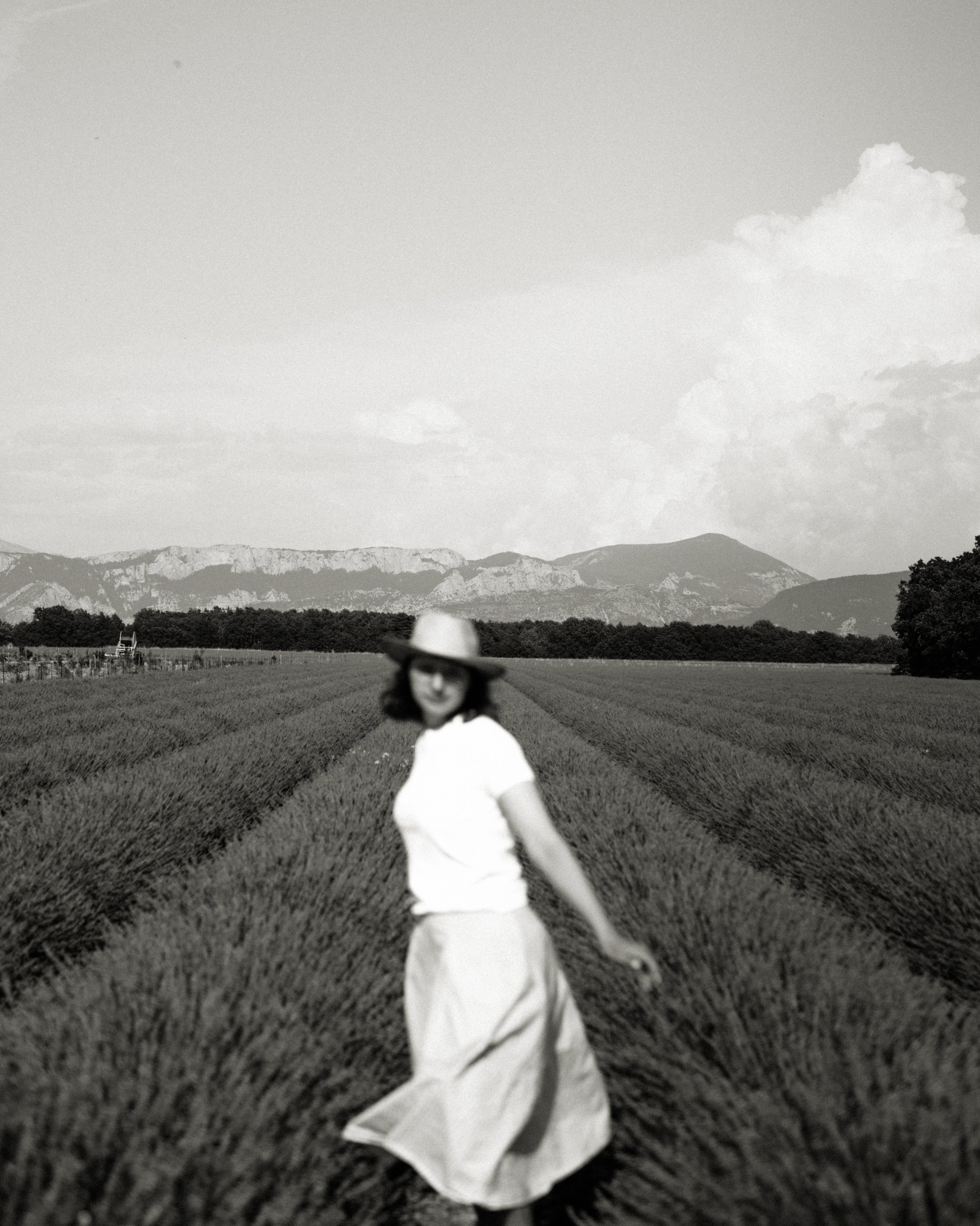A woman in a white blouse, wide-brimmed hat, and long skirt walking through a cultivated field with mountains in the background, black and white photo.