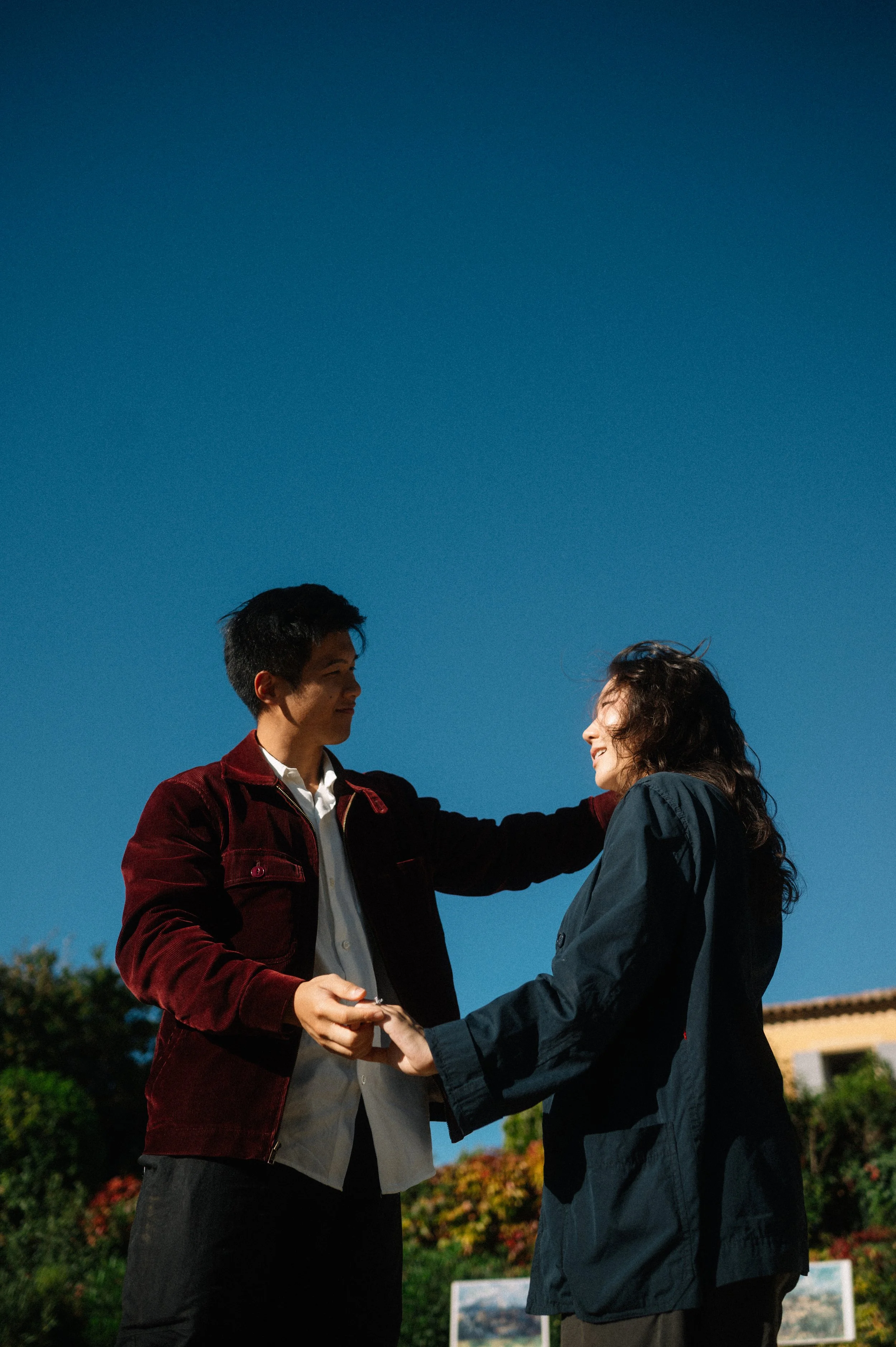 A young man and woman holding hands and smiling at each other outdoors against a clear blue sky, with plants and a building in the background.