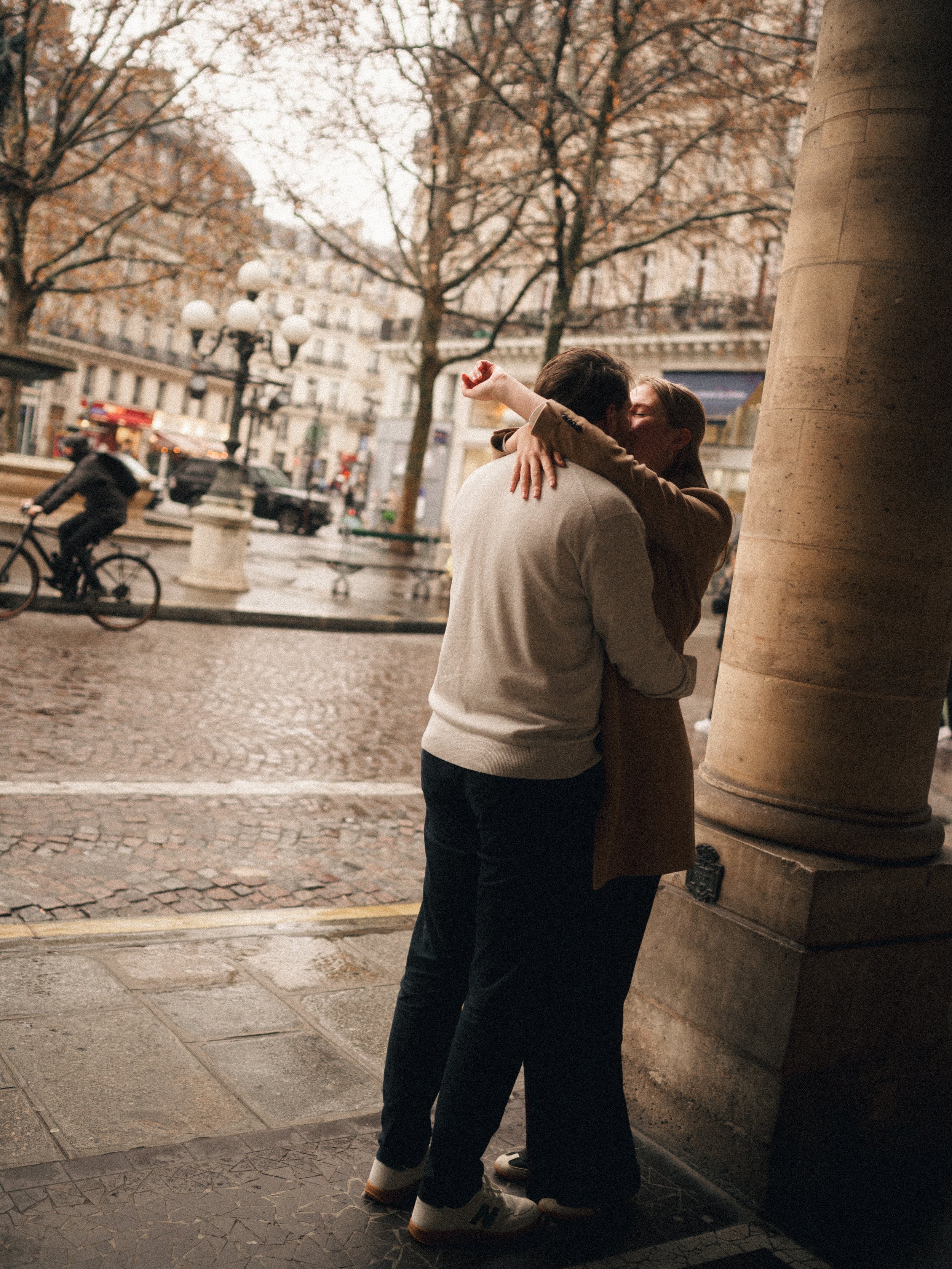 A couple embracing each other under a street overhang in an urban setting on a rainy day, with a cyclist and buildings in the background.