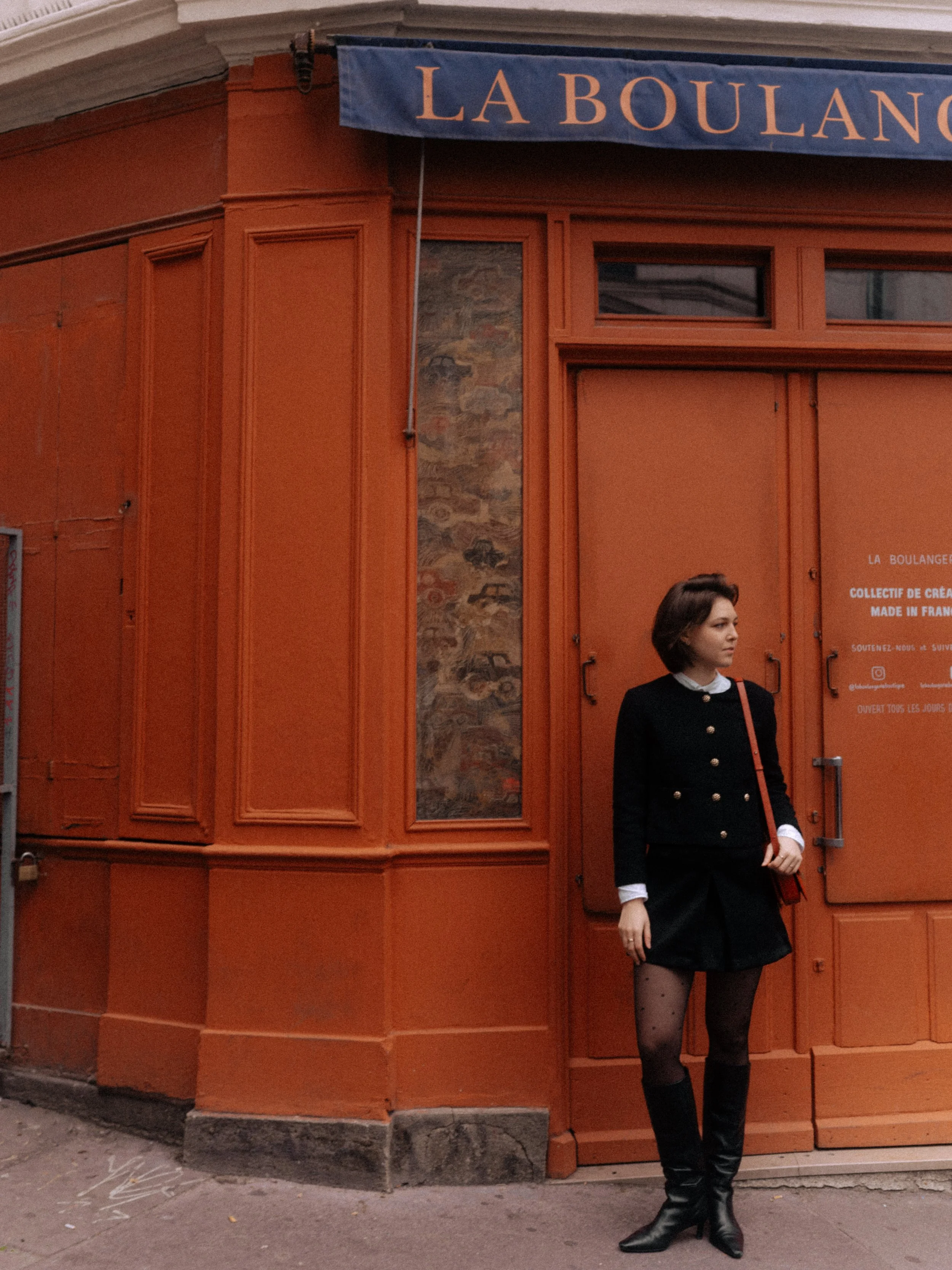 A woman with short brown hair wearing a black coat, skirt, patterned tights, and knee-high black boots standing in front of an orange-colored storefront with a blue sign that reads "LA BOULANGERIE."