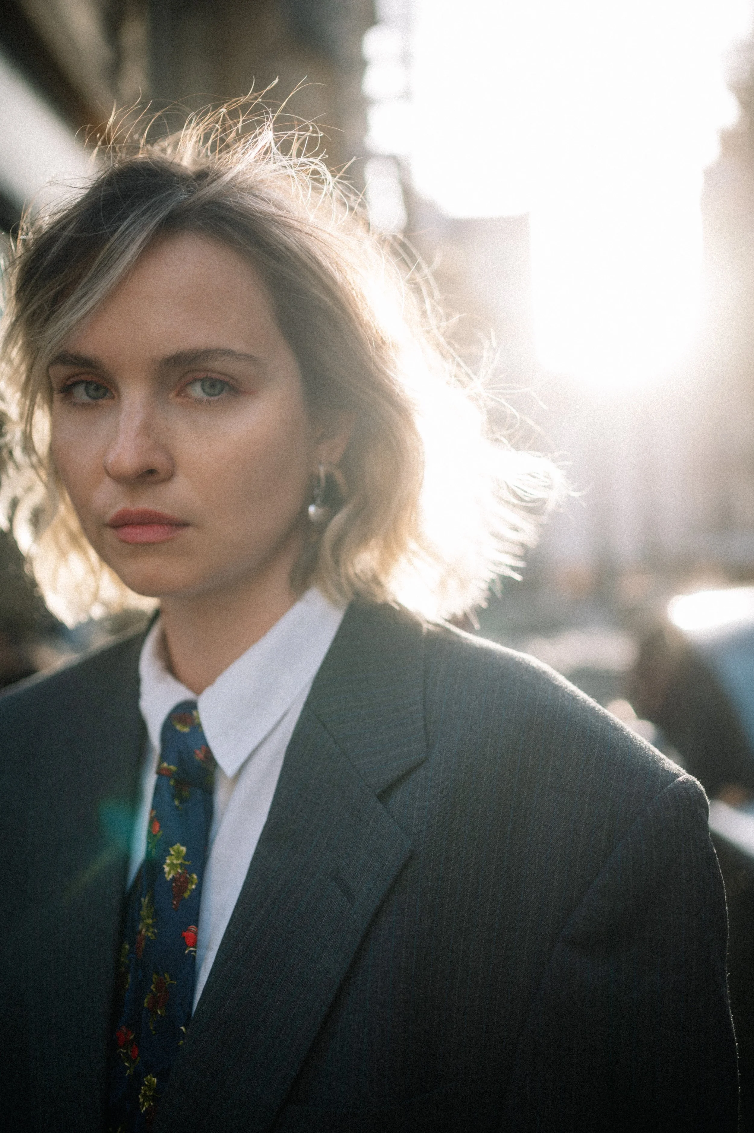 A woman with blonde, wavy hair wearing a dark suit, white shirt, and floral tie, standing outdoors in sunlight during sunset.